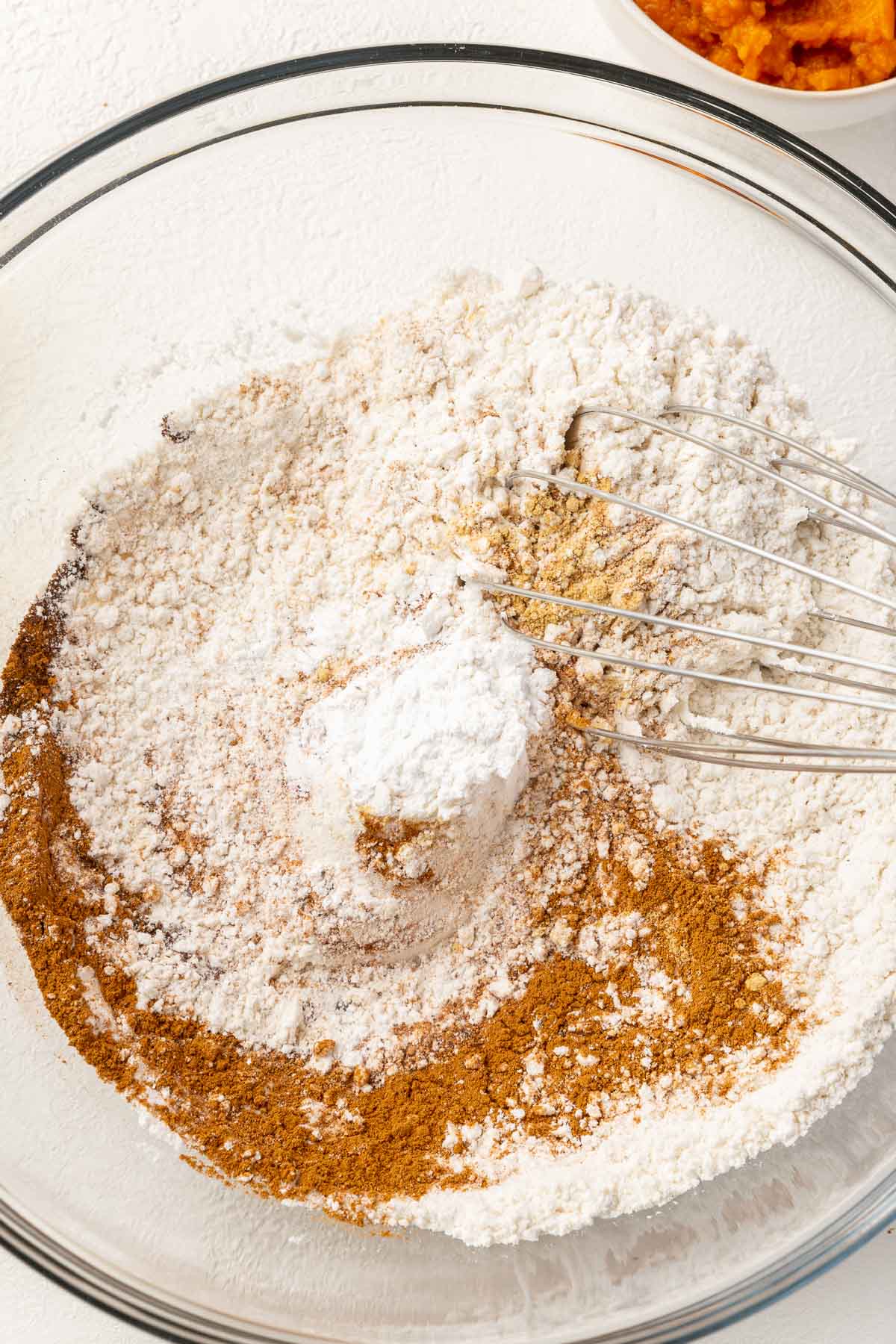 A glass mixing bowl with gluten-free flour, cinnamon, baking soda, baking powder, ginger, cloves, and nutmeg being mixed together with a whisk.