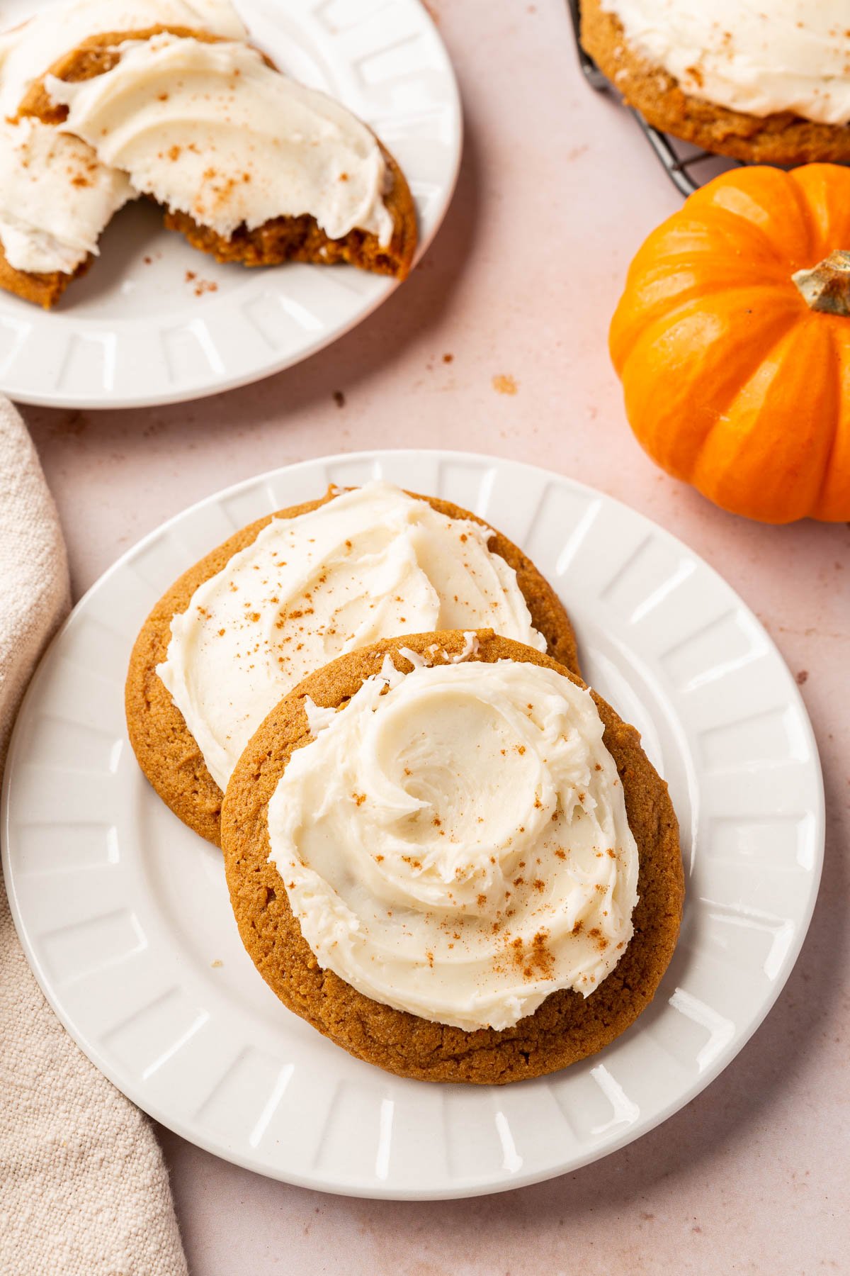 Two gluten-free pumpkin cookies with cream cheese frosting on a dessert plate with additional cookies in the background.