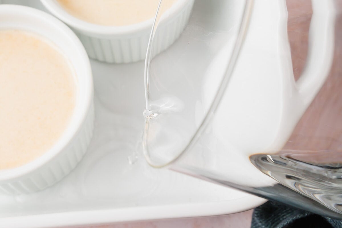 A glass measuring cup pouring water into a bain marie around ramekins filled with pumpkin crème brûlée mixture.
