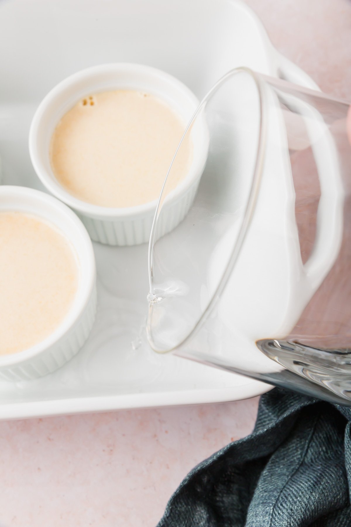 A glass measuring cup pouring water into a bain marie around ramekins filled with pumpkin crème brûlée mixture.