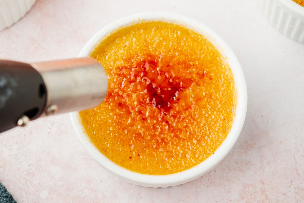A kitchen torch caramelizing granulated sugar on top of pumpkin crème brûlée in a ramekin.