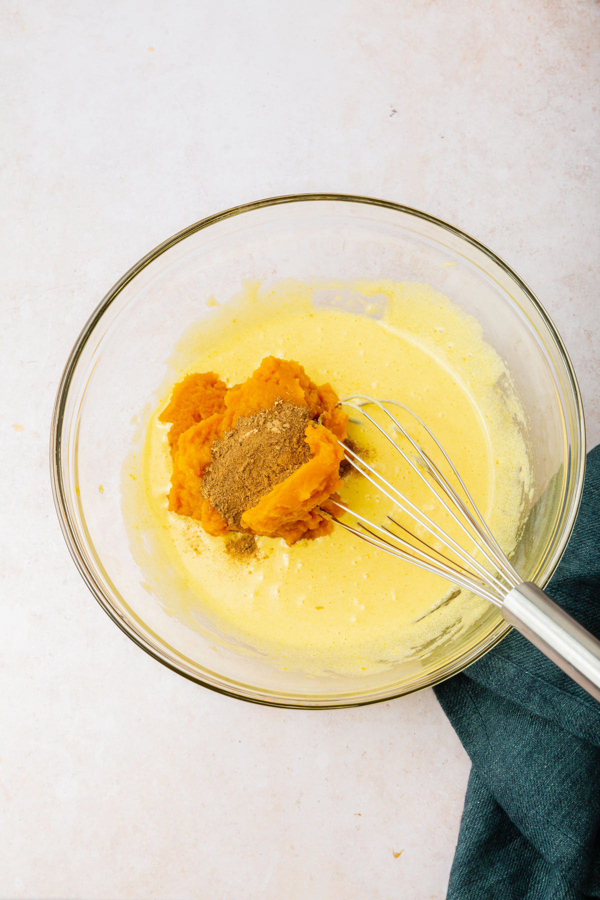 A glass mixing bowl with a light yellow egg yolk and sugar mixture topped with pumpkin puree and pumpkin pie spice before mixing together.