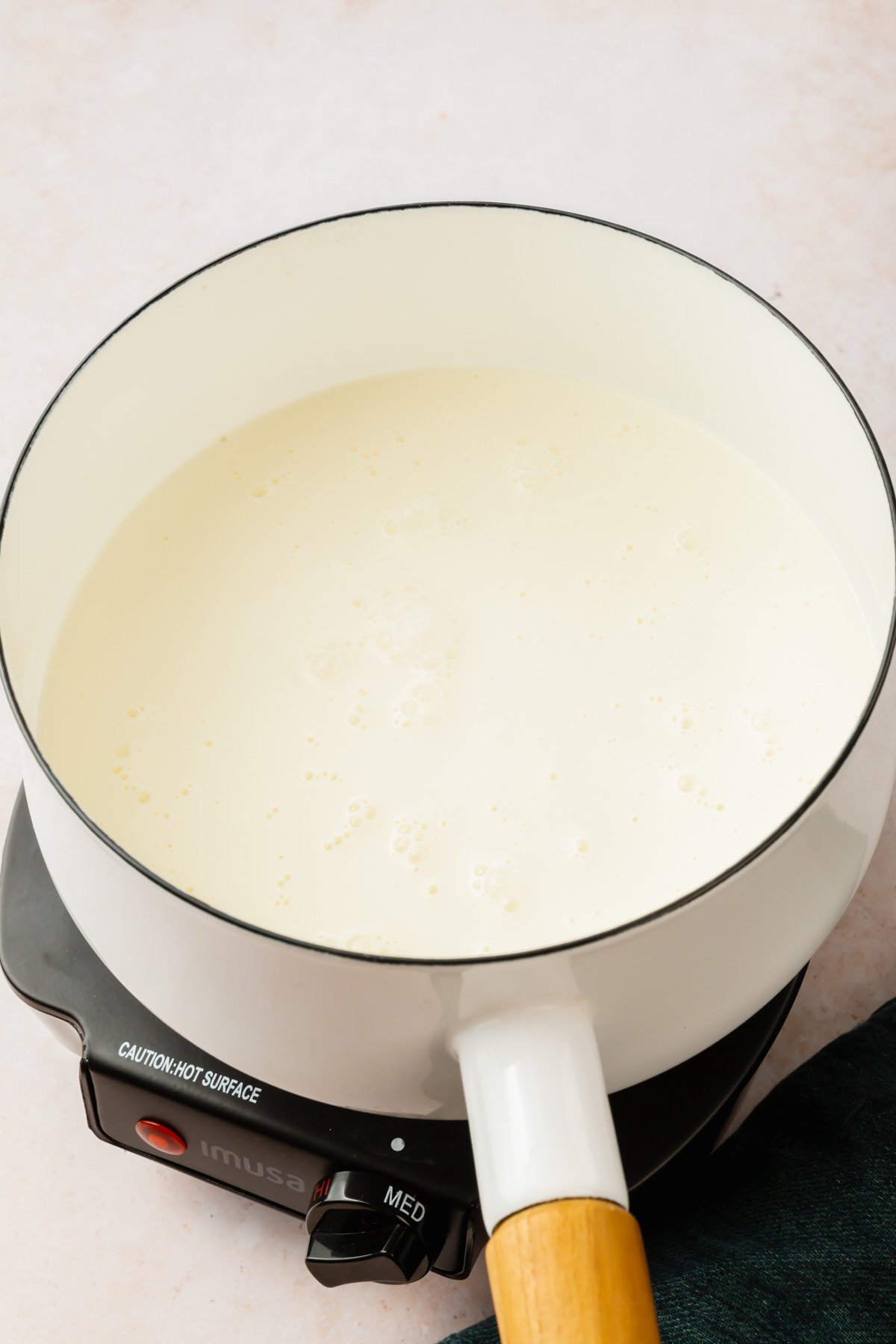A white saucepan on a portable burner with heavy cream in it that is simmering.