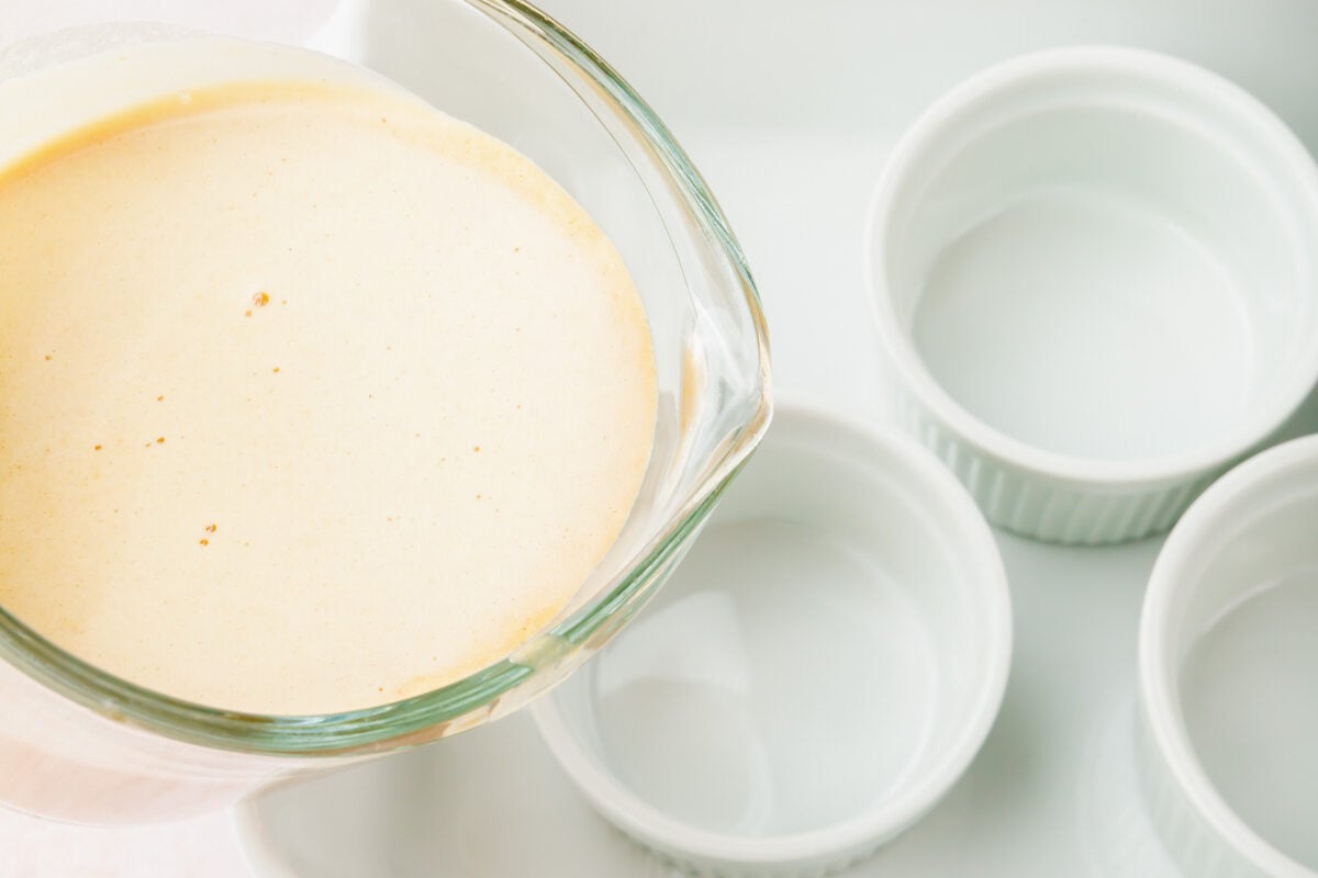 A glass measuring cup pouring pumpkin crème brûlée mixture into individual ramekins in a ceramic baking pan.