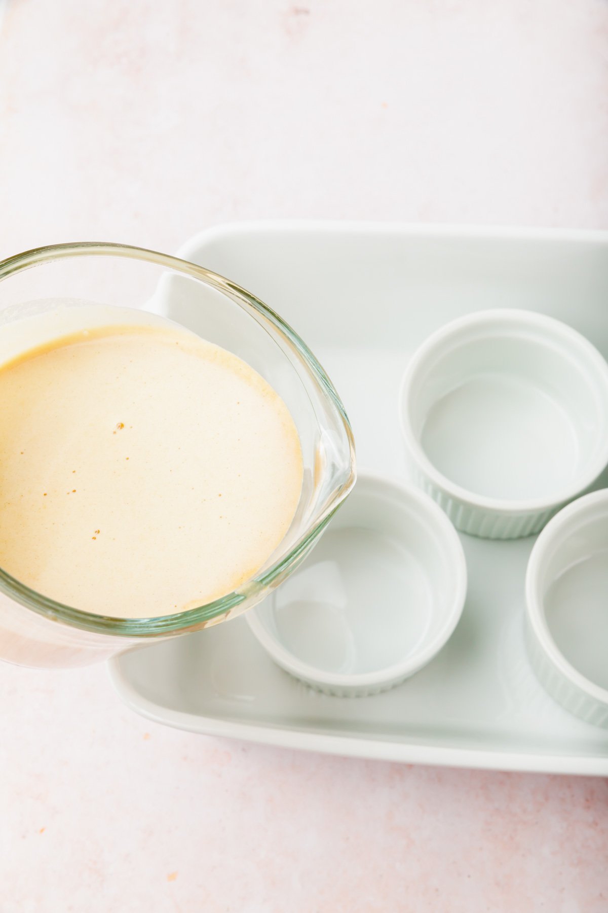 A glass measuring cup pouring pumpkin crème brûlée mixture into individual ramekins in a ceramic baking pan.