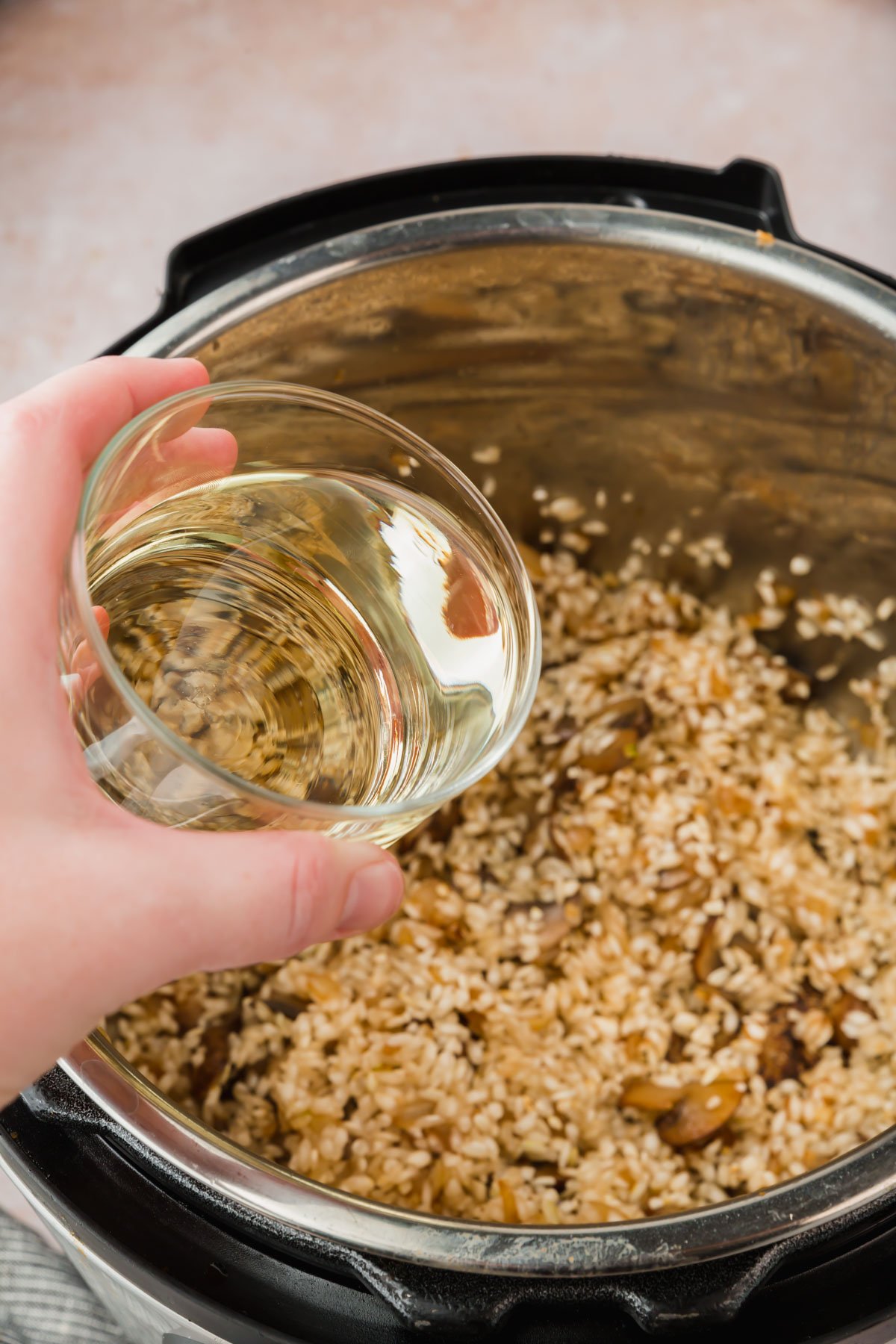 A hand pouring a glass of dry white wine into an Instant Pot filled with arborio rice and mushrooms.