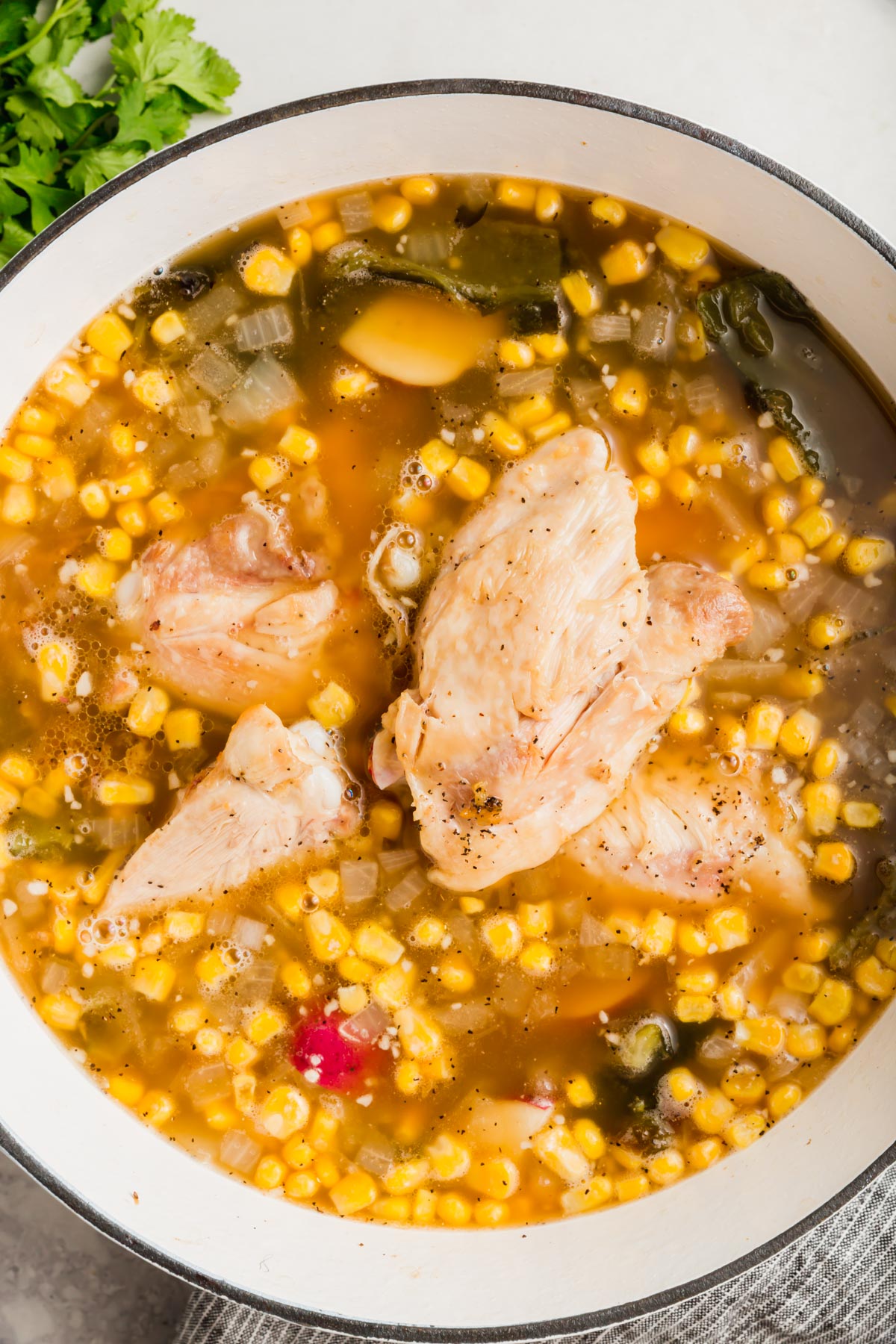 An overhead view of a white dutch oven with poblano corn soup topped with cooked chicken thighs.