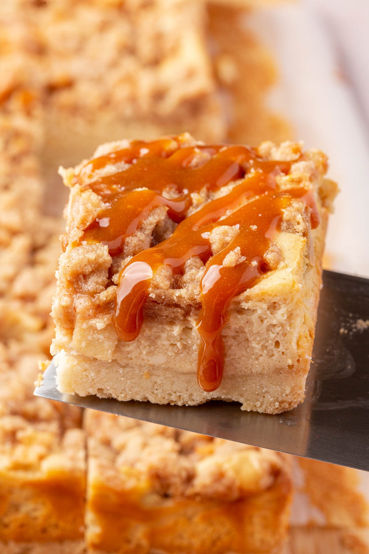 A metal spatula lifting a gluten free apple caramel cheesecake bar with streusel out of a pan of additional gluten free cheesecake bars.