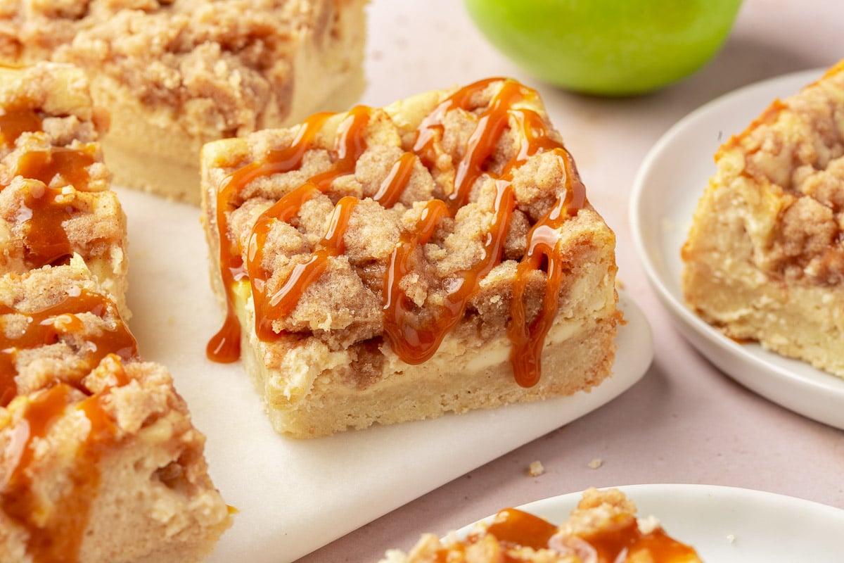 A gluten free caramel apple cheesecake bar on a board drizzled with caramel sauce with additional bars in the background and a green apple.