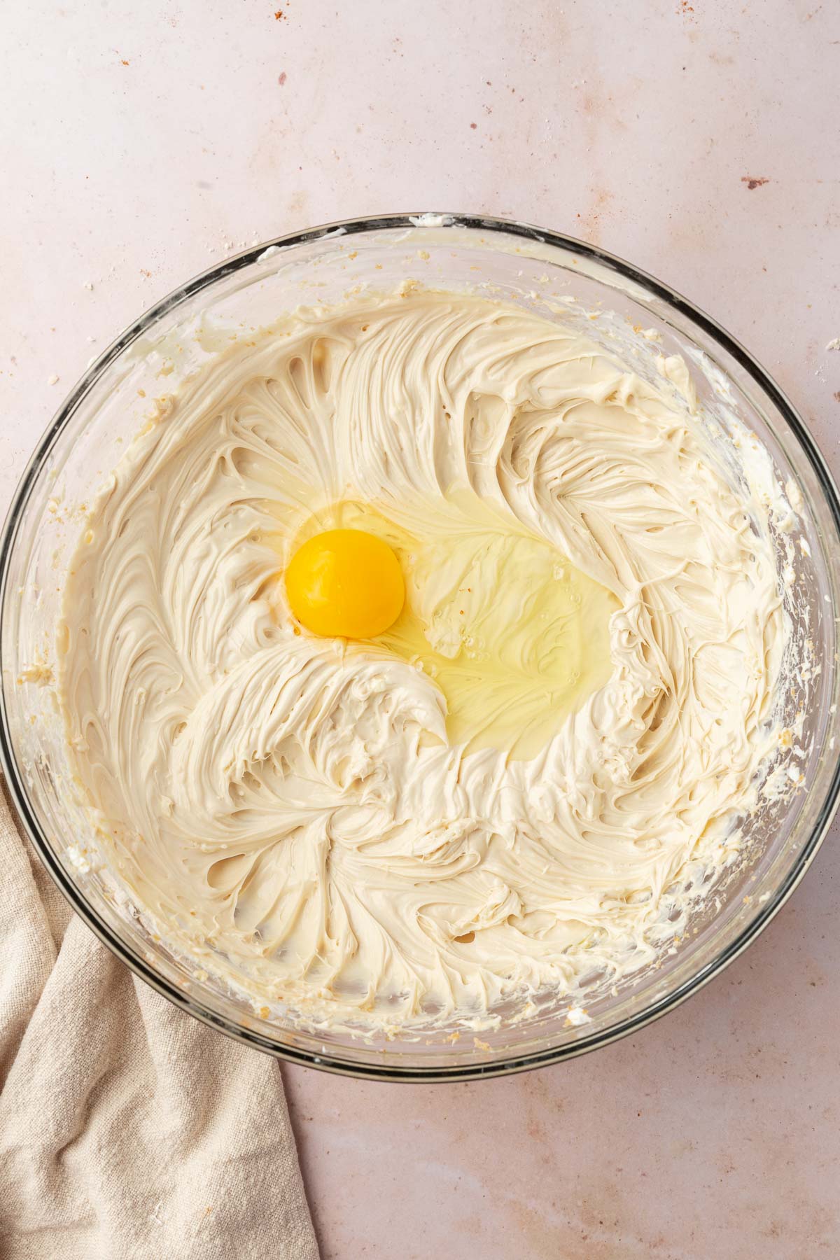 A glass mixing bowl with a cheesecake filling mixture topped with one egg before mixing together.