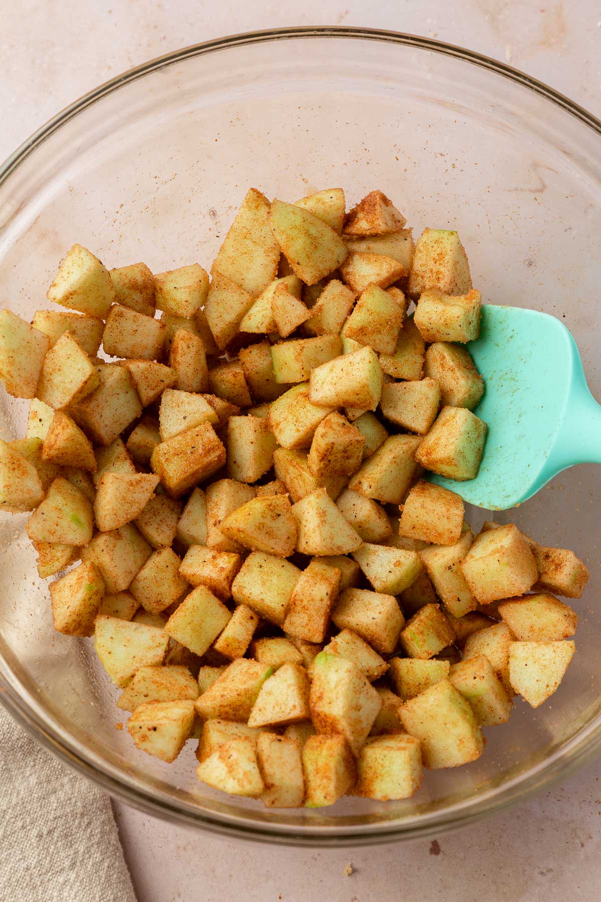 A glass mixing bowl with diced granny smith apples in it that has been mixed together with ground cinnamon and brown sugar with a blue rubber spatula.