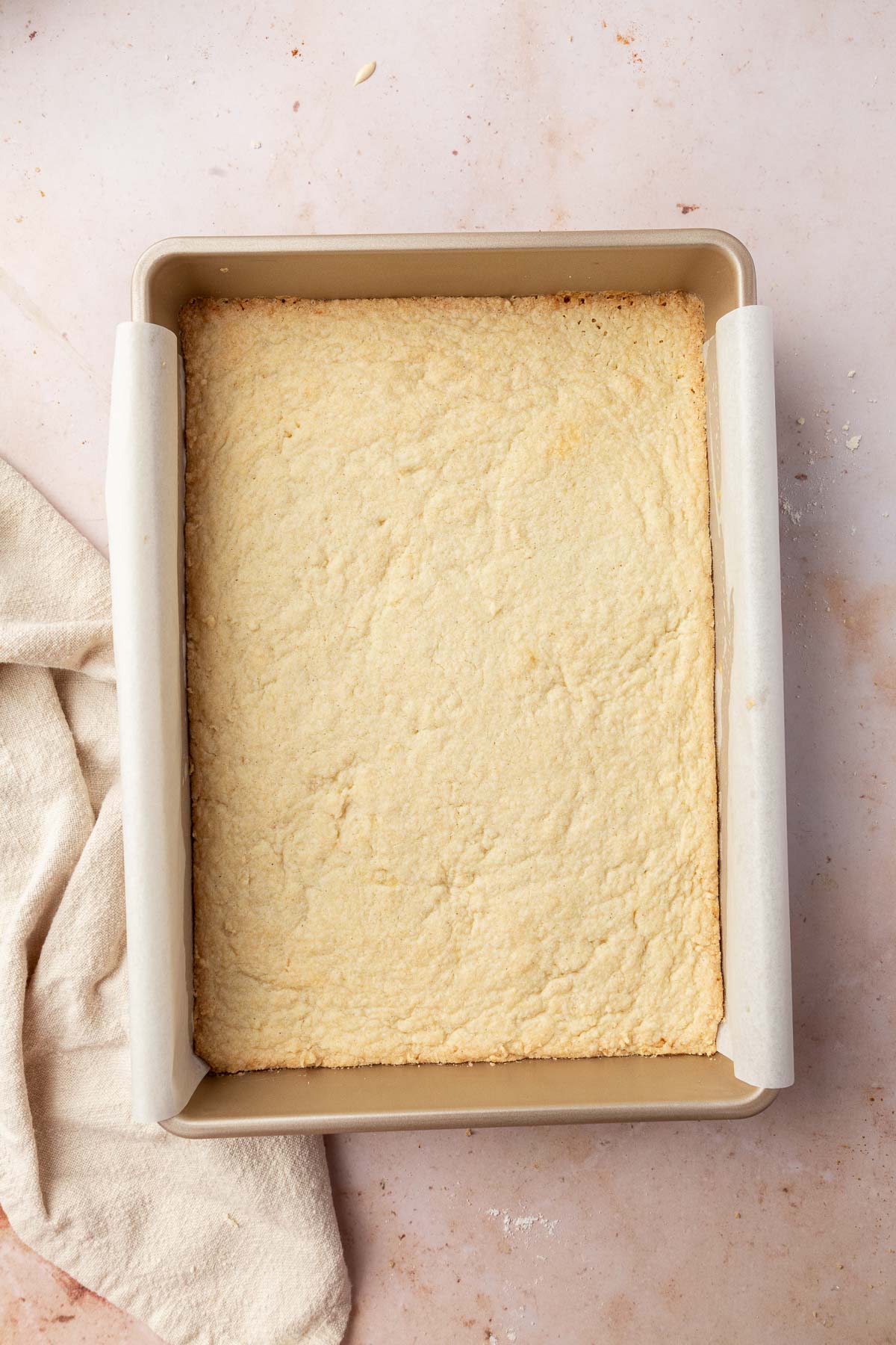 A gold metal rectangular baking pan lined with parchment paper and filled with a golden baked gluten-free shortbread crust.