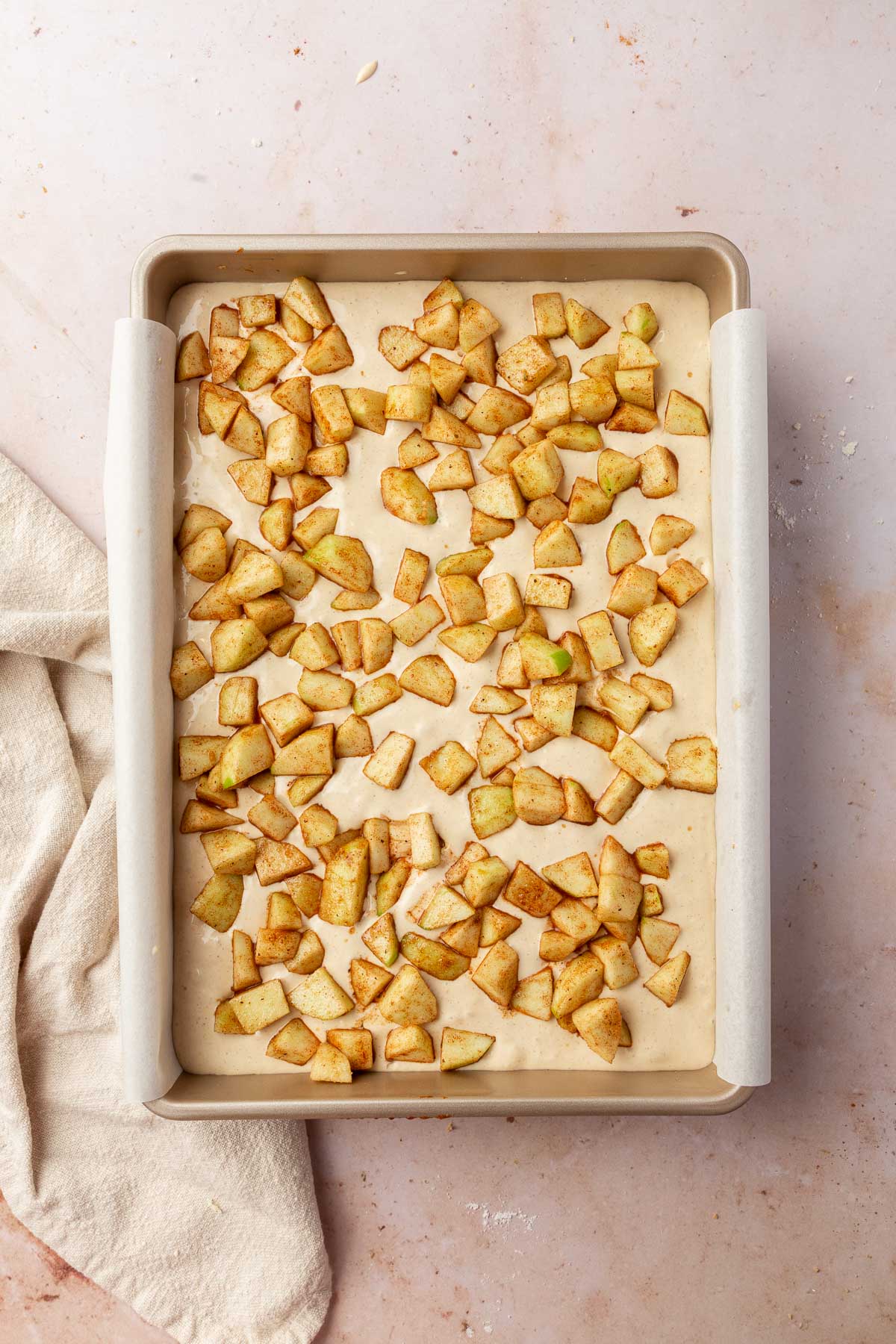 A rectangular baking pan filled with a cheesecake mixture topped with cinnamon spiced apple chunks before baking in the oven.