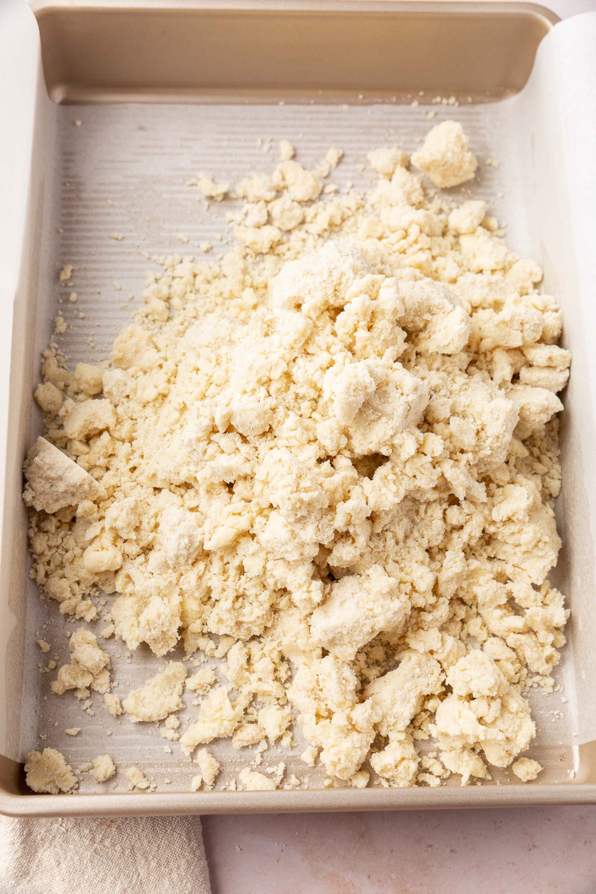 A parchment lined metal baking pan that has a pile of gluten-free shaggy dough to make a shortbread crust layer for cheesecake.