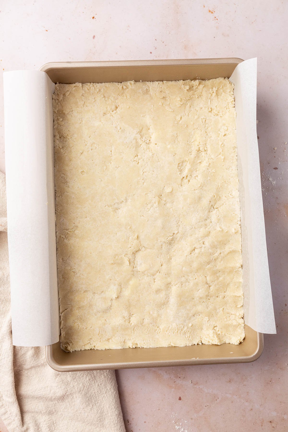 An overhead view of a rectangular baking pan lined with parchment paper that has a gluten-free shortbread crust pressed into it before baking in the oven.