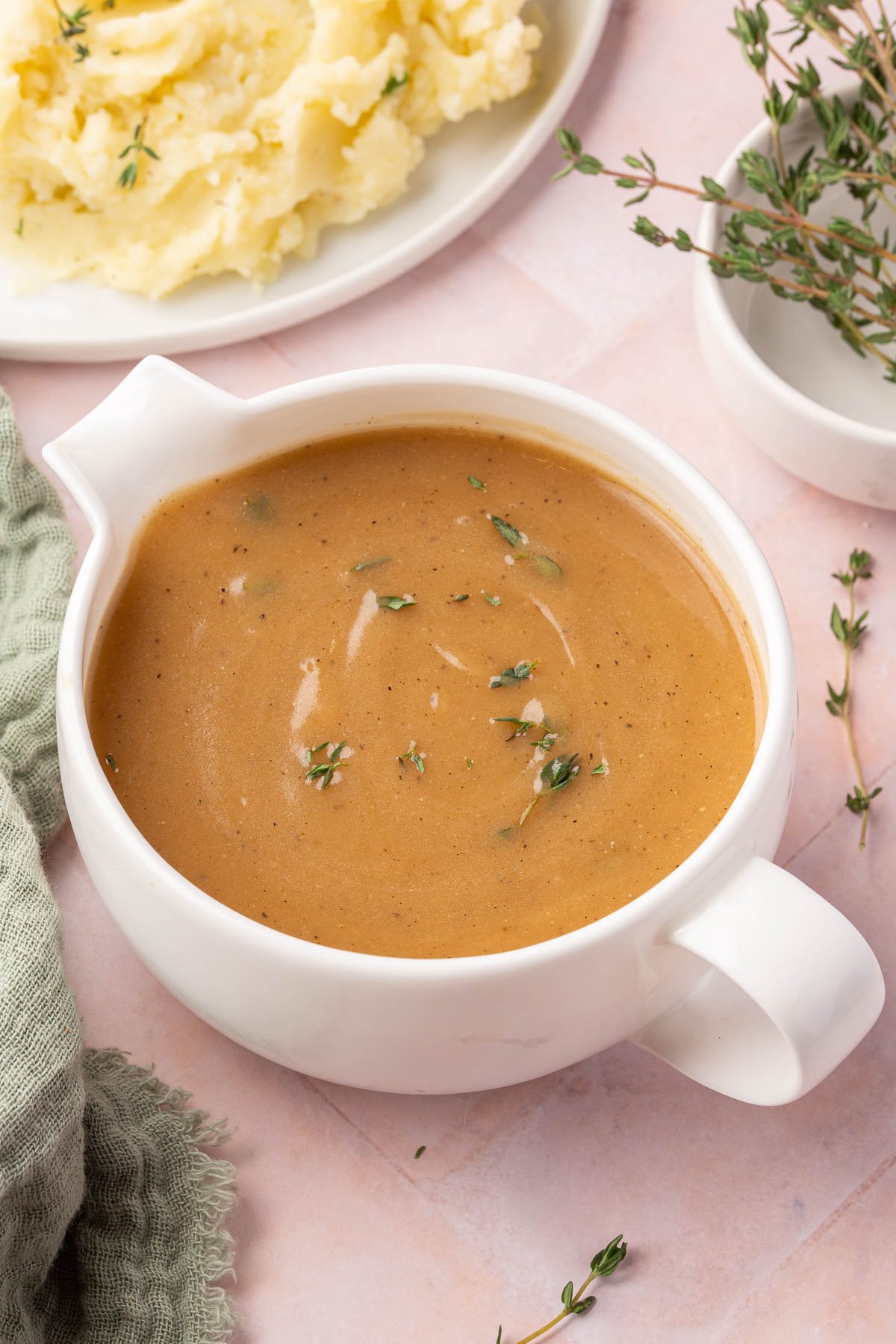 A round gravy boat filled with gluten-free gravy topped with fresh thyme with a serving of mashed potatoes in the corner.