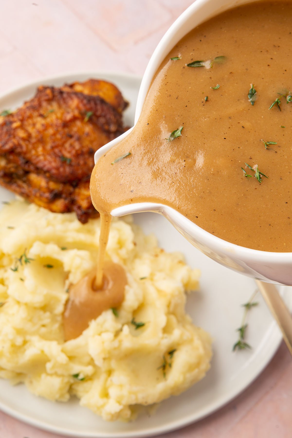 A gravy boat pouring gluten-free gravy over a serving of mashed potatoes with a roasted chicken thigh in the background.