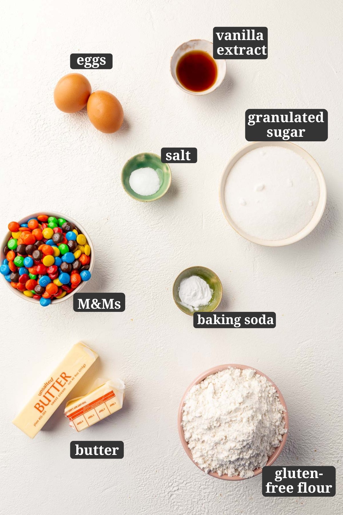 Ingredients in small bowls to make gluten-free M&M sugar cookies, including two eggs, vanilla extract, salt, granulated sugar, M&Ms, baking soda, butter and gluten-free flour with text overlays over each ingredient.