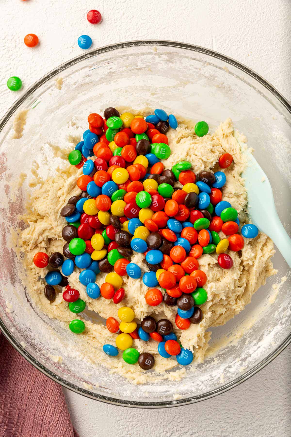 A glass mixing bowl with gluten-free sugar cookie dough topped with M&Ms before mixing together. I