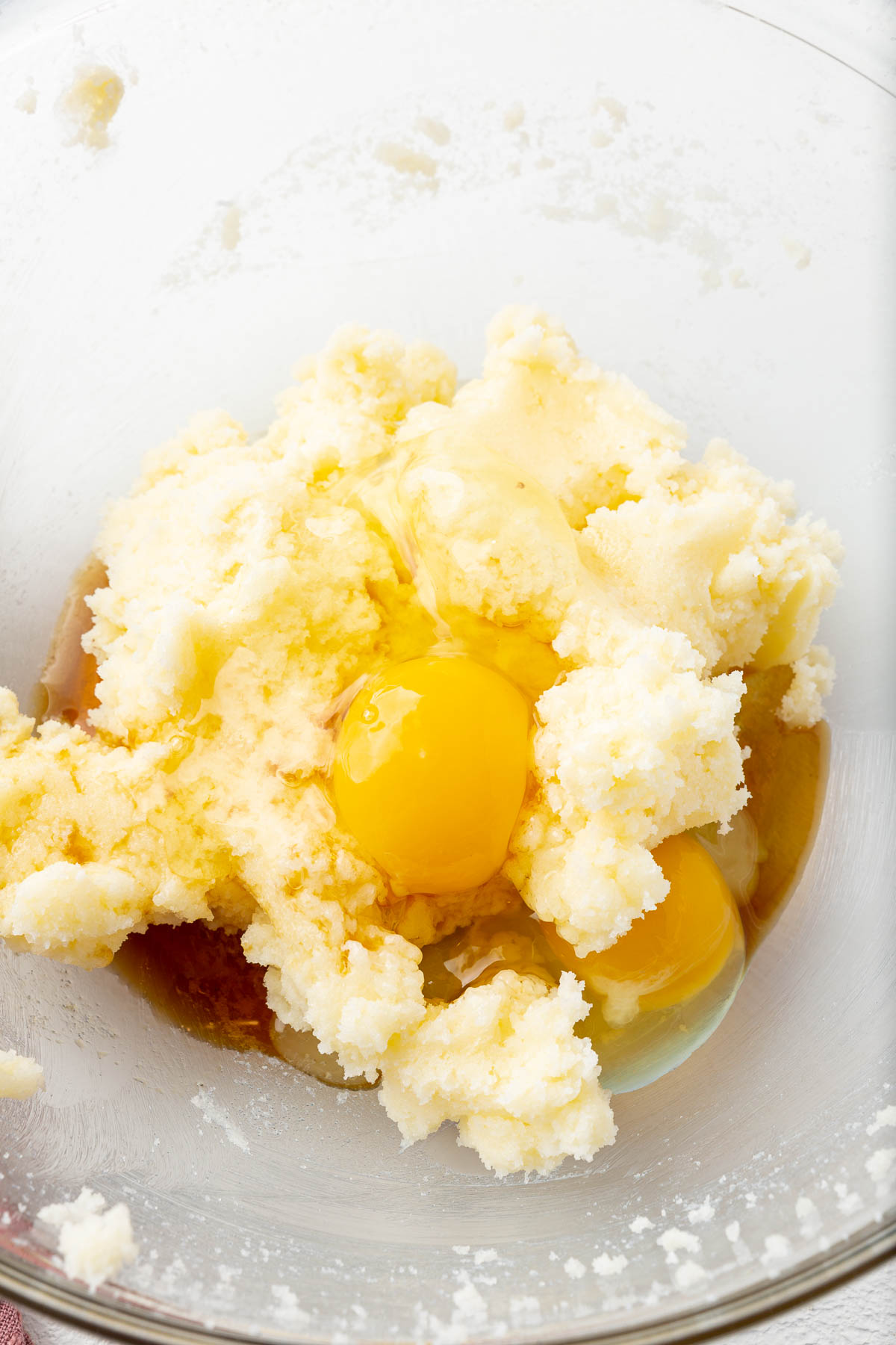 A close up of a glass mixing bowl with creamed butter and granulated sugar topped with two eggs and vanilla extract before mixing together.