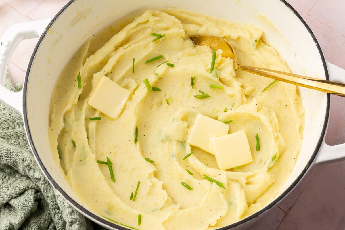 A white dutch oven with mashed potatoes topped with three pats of butter and chopped chives with a gold serving fork.