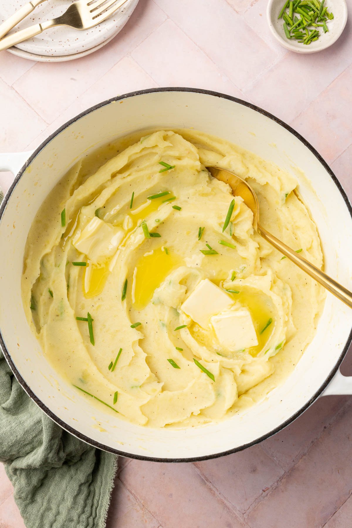 An overhead view of a dutch oven with mashed potatoes and pats of melted butter and freshly chopped chives with a gold serving spoon.