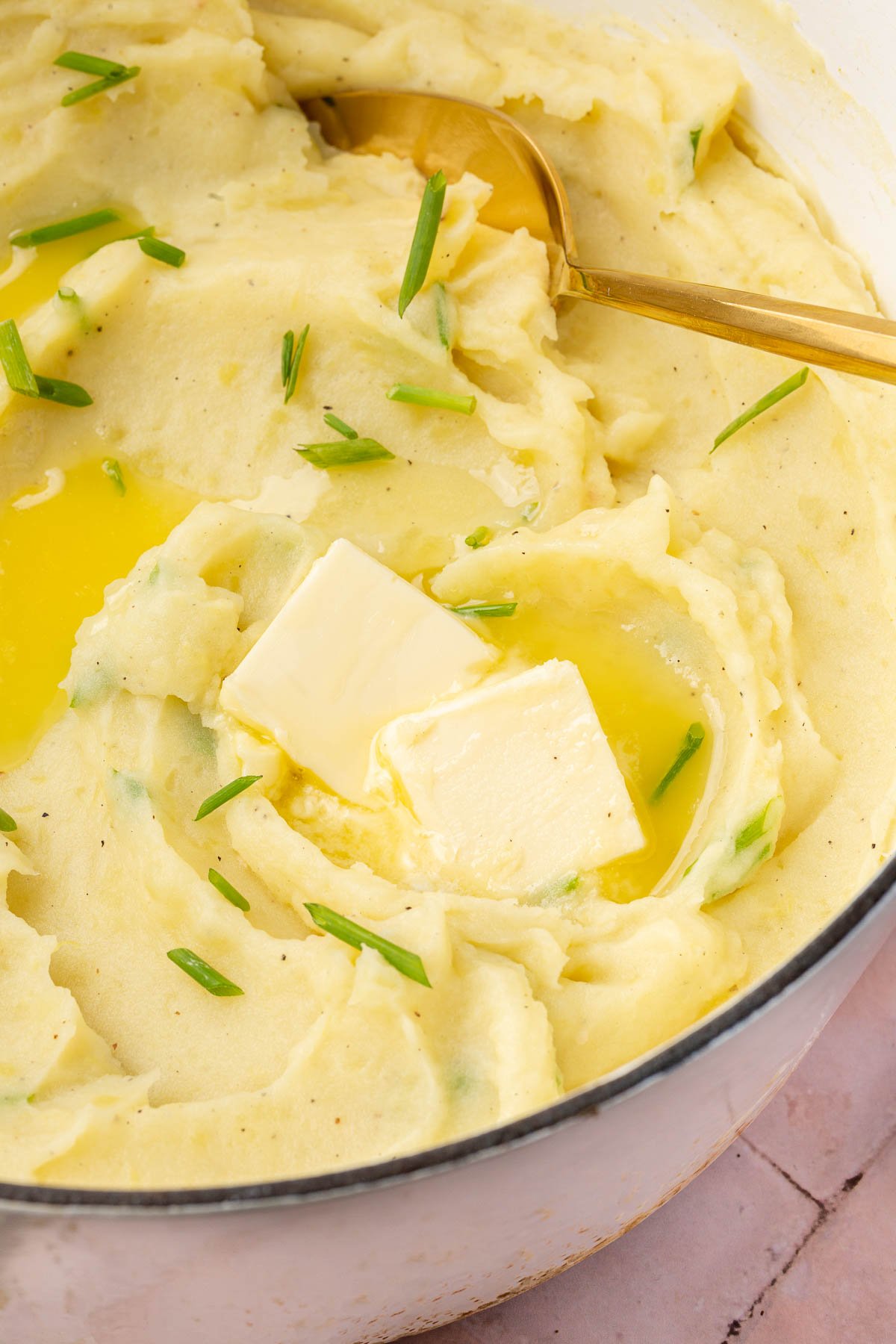 A white dutch oven of gluten-free mashed potatoes with two pats of melted butter and fresh chopped chives with a gold serving spoon.