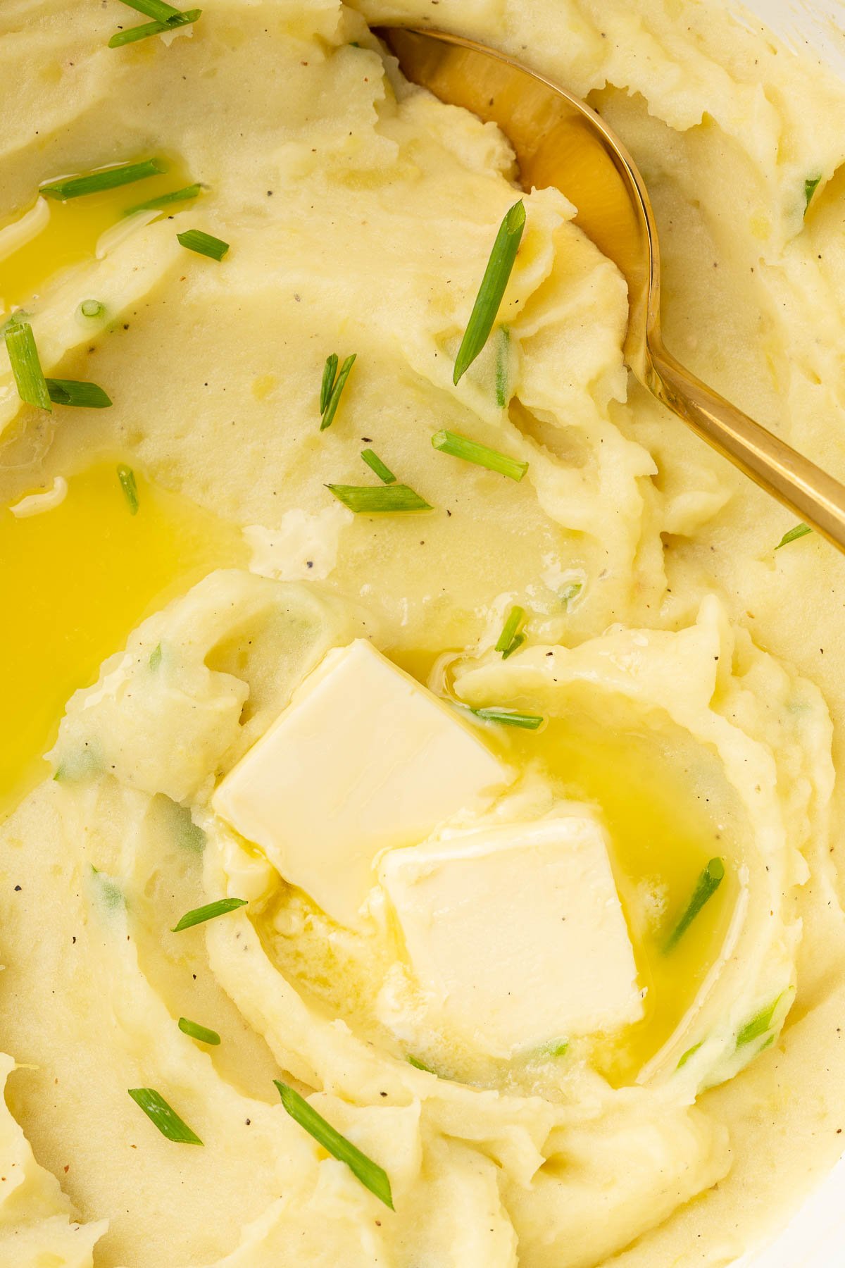 A close up of gluten-free mashed potatoes in a dutch oven with pats of melted butter and fresh chives with a gold serving spoon.