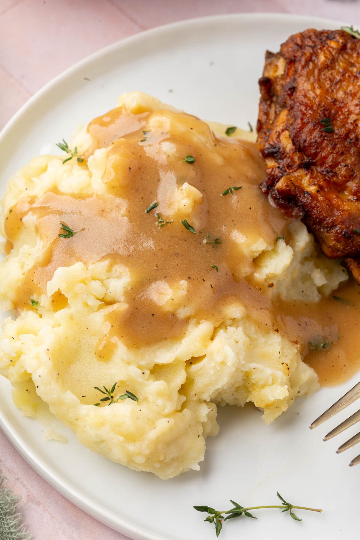 A serving of mashed potatoes topped with gravy and fresh thyme with crispy chicken thighs in the background.