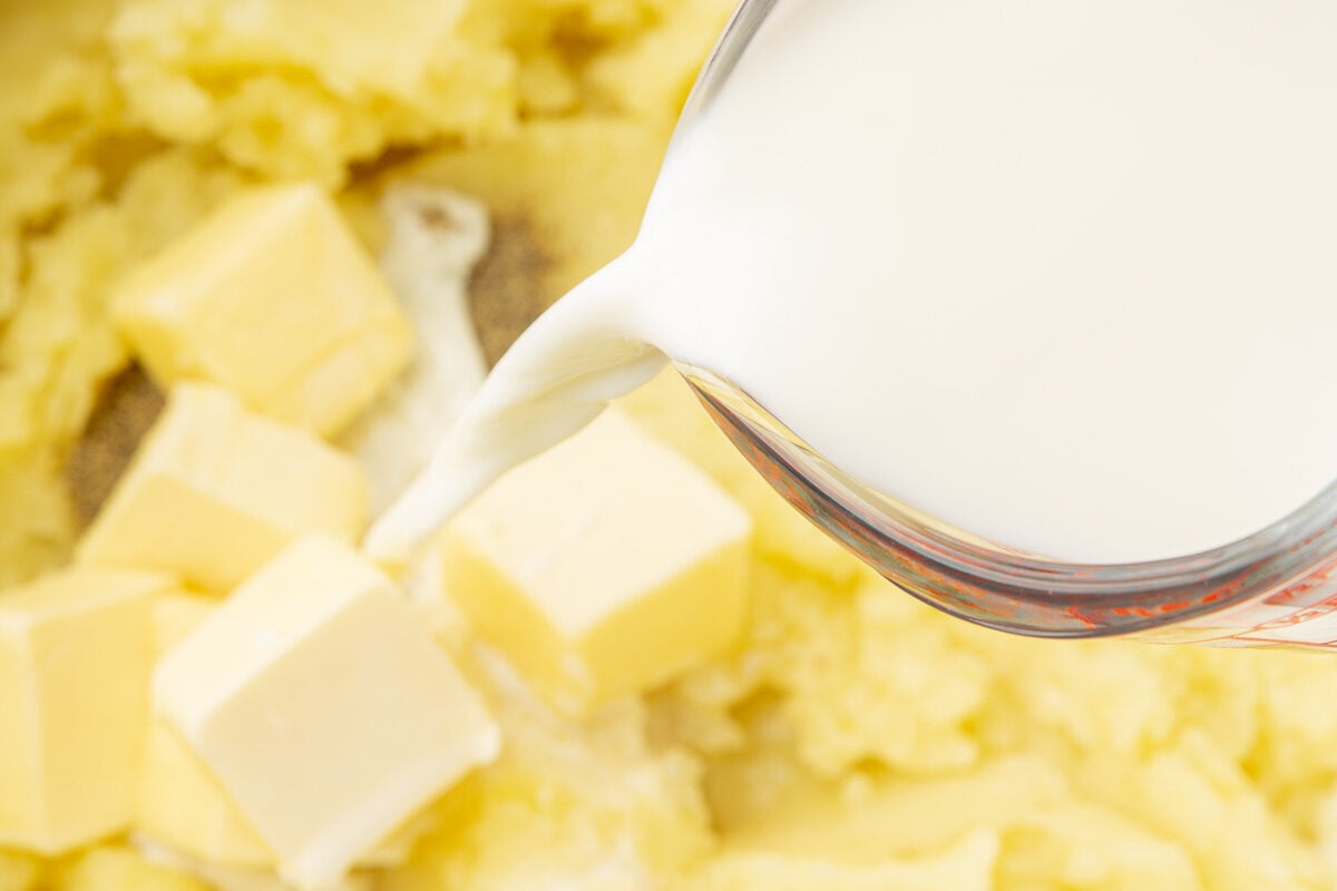 A glass measuring cup of milk being poured into a dutch oven of mashed potatoes with butter.