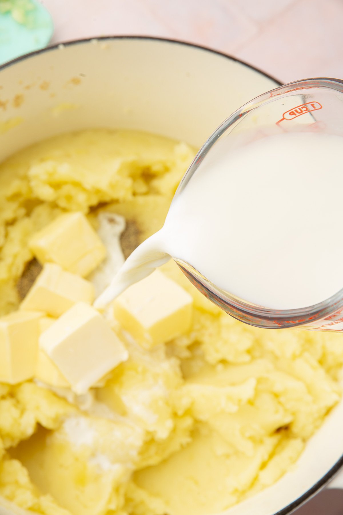 A glass measuring cup of milk being poured into a dutch oven of mashed potatoes with butter.