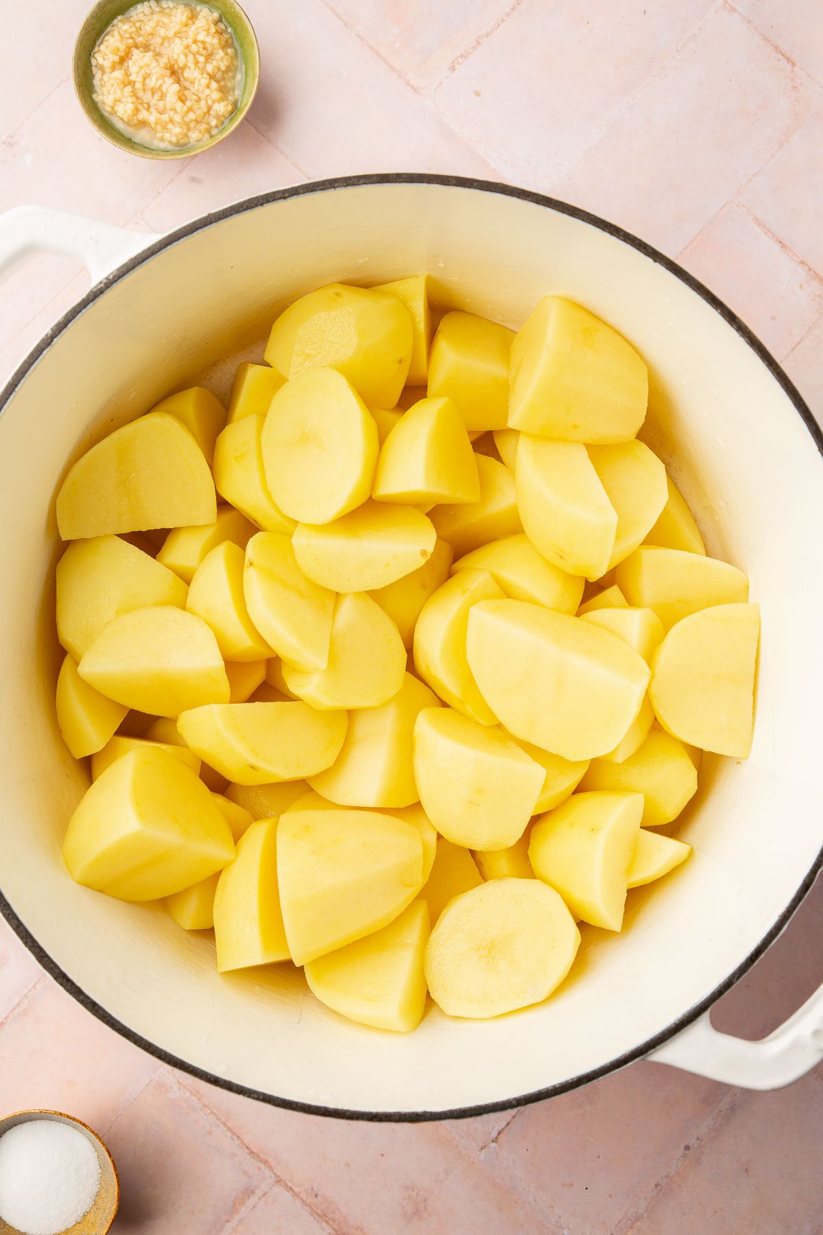 A pot of peeled quartered potatoes with a bowl of garlic and salt on the side.