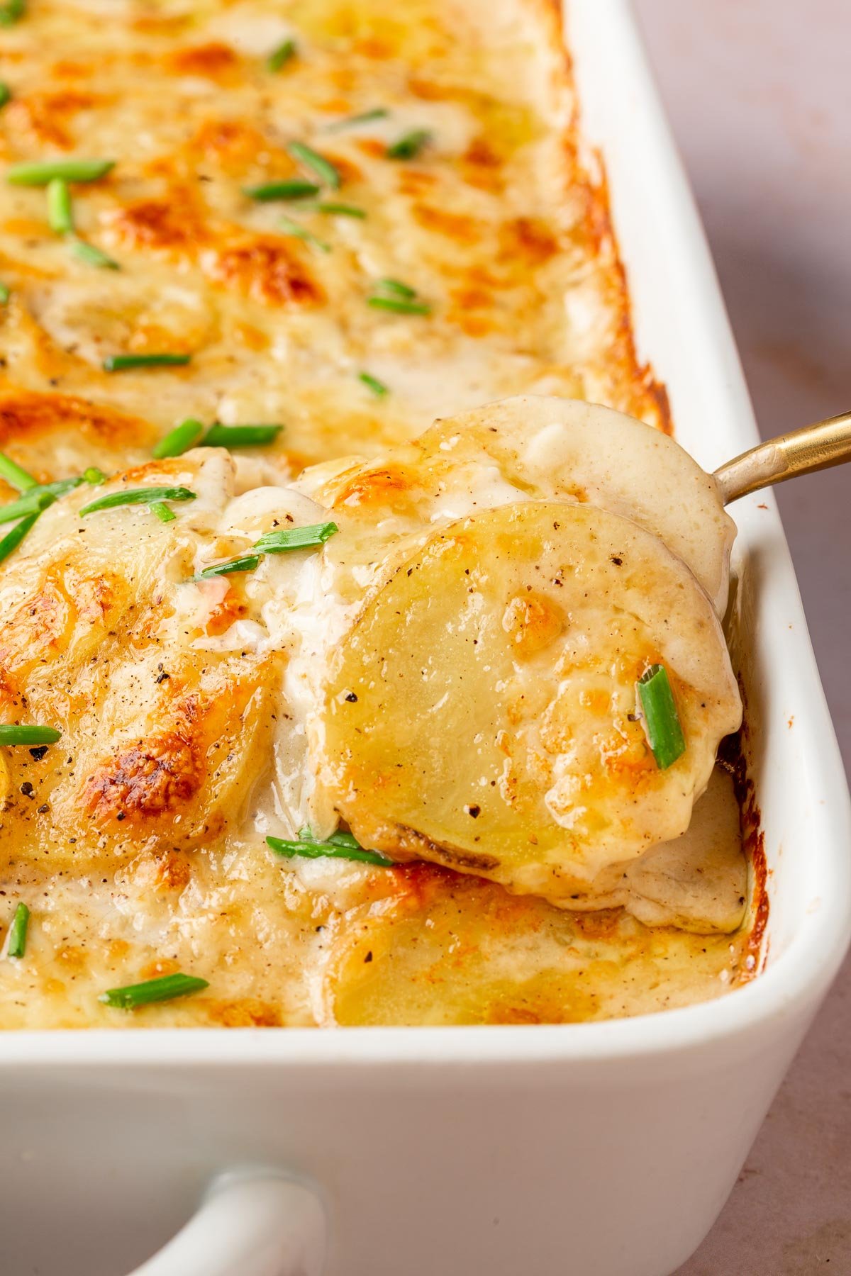 A gold serving spoon scooping gluten-free scalloped potatoes out of a white rectangle baking dish.