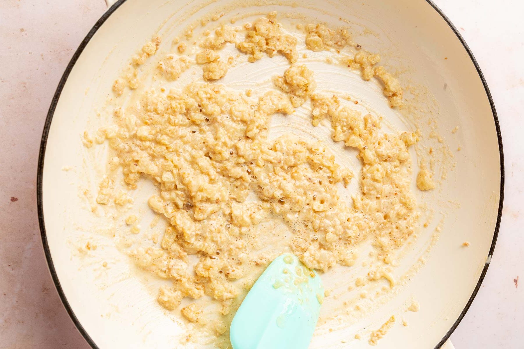 A braising pan with melted butter, sautéed onions and garlic mixed together with gluten-free flour that is being stirred together with a blue spatula to make a gluten-free roux.