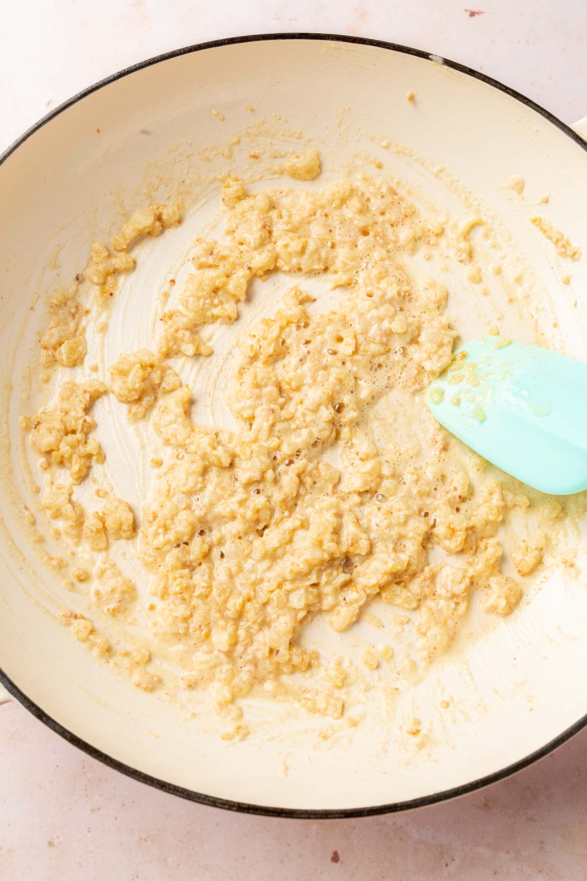 A braising pan with melted butter, sautéed onions and garlic mixed together with gluten-free flour that is being stirred together with a blue spatula to make a gluten-free roux.
