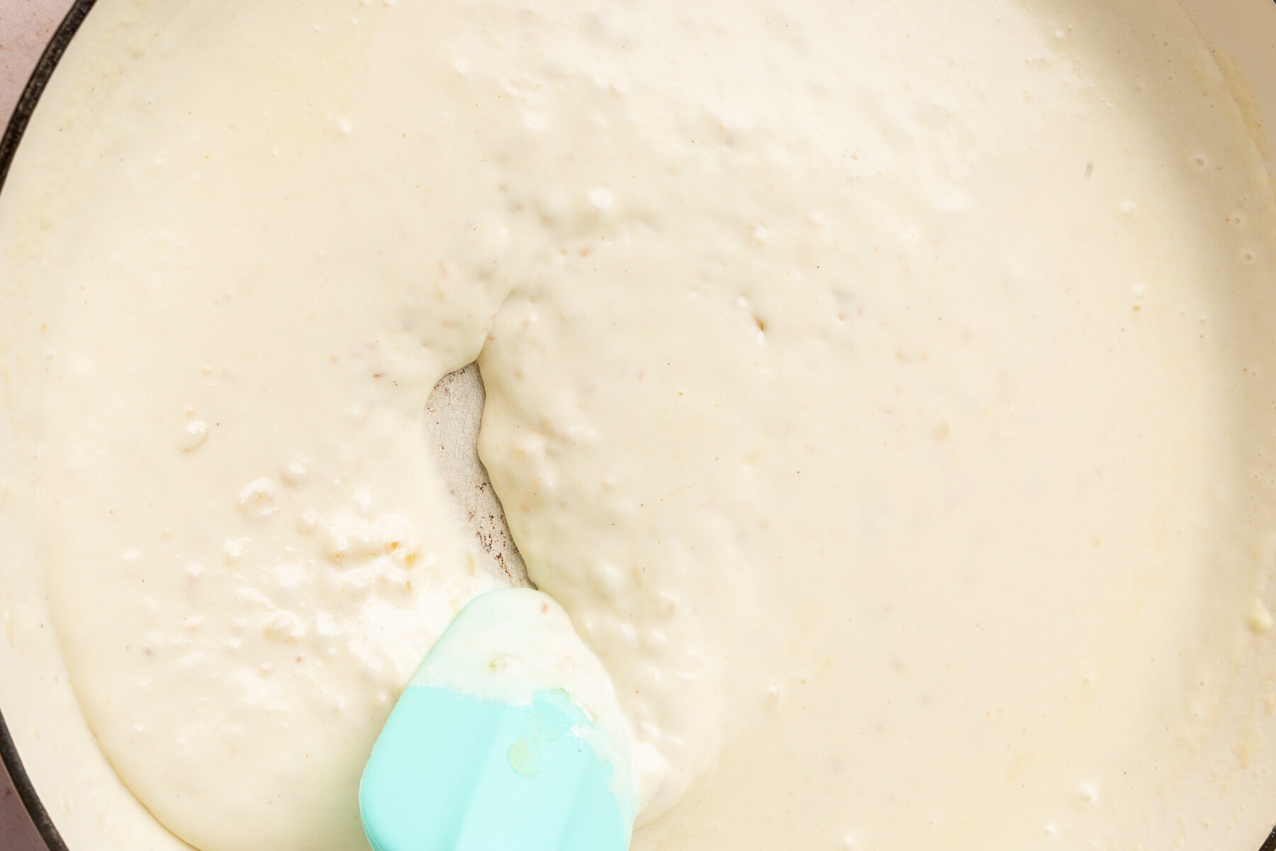 A braising pan with a thick cream sauce being stirred with a blue rubber spatula.