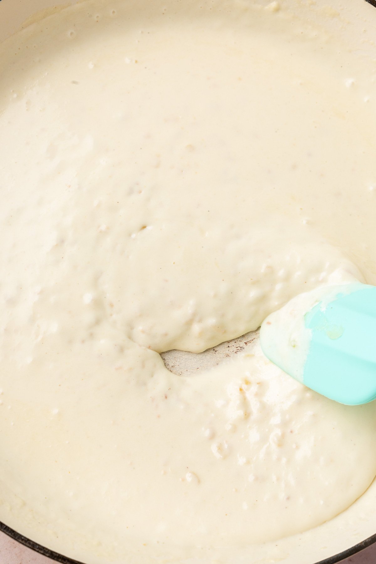 A braising pan with a thick cream sauce being stirred with a blue rubber spatula.