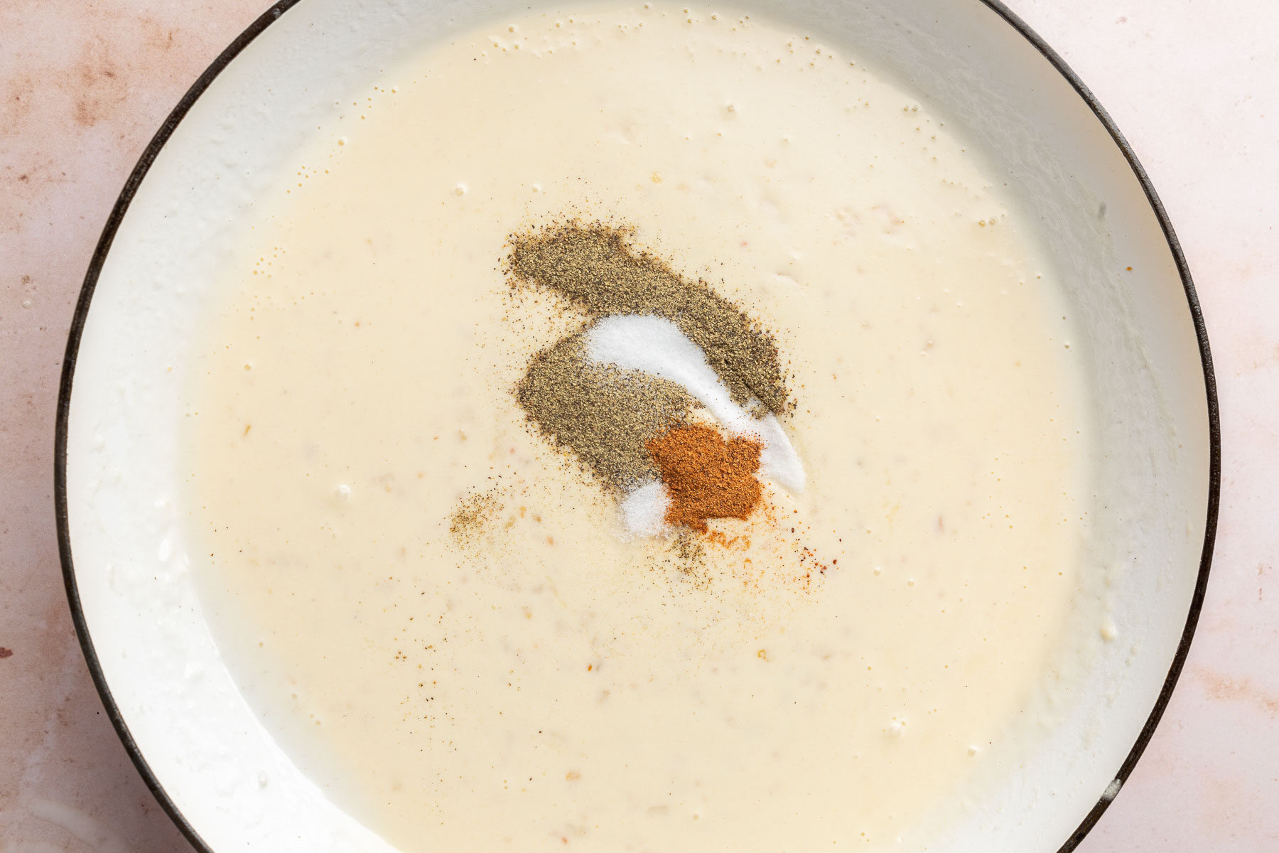 A braising pan with a thick gluten-free cream sauce topped with black pepper, salt, and nutmeg before stirring together.
