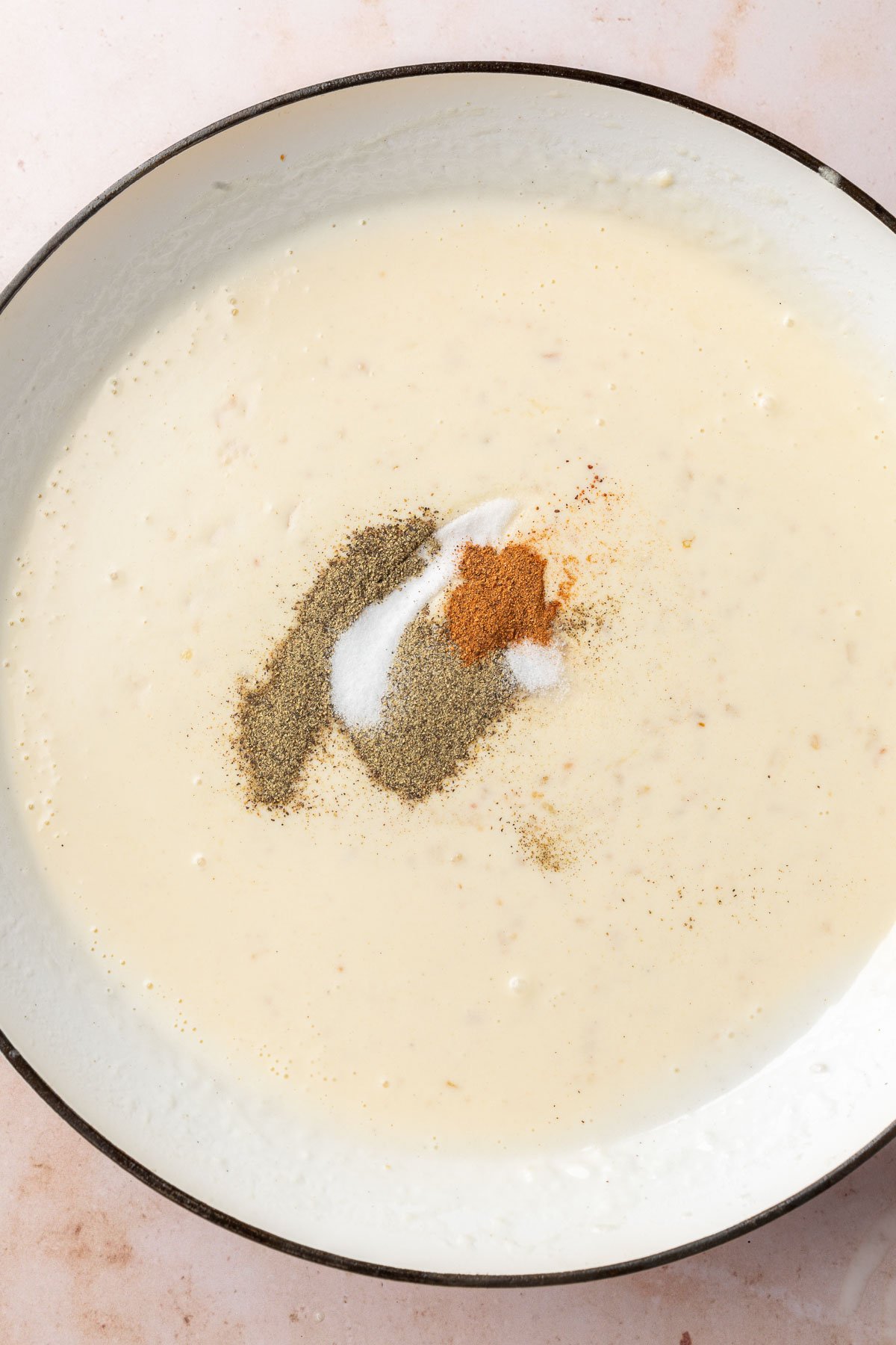 A braising pan with a thick gluten-free cream sauce topped with black pepper, salt, and nutmeg before stirring together.