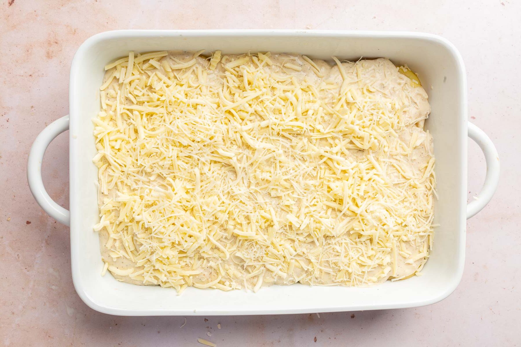 A white rectangular baking dish with a gluten-free cream sauce and shredded white cheddar and parmesan cheeses.