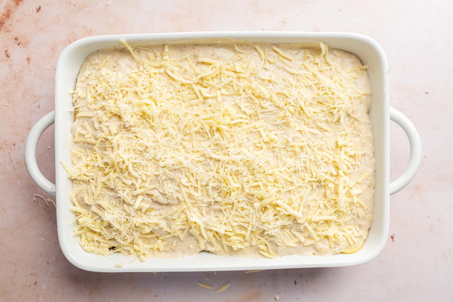 A rectangular baking dish with layers of gluten-free cream sauce topped with shredded white cheddar cheese and shredded parmesan cheese.