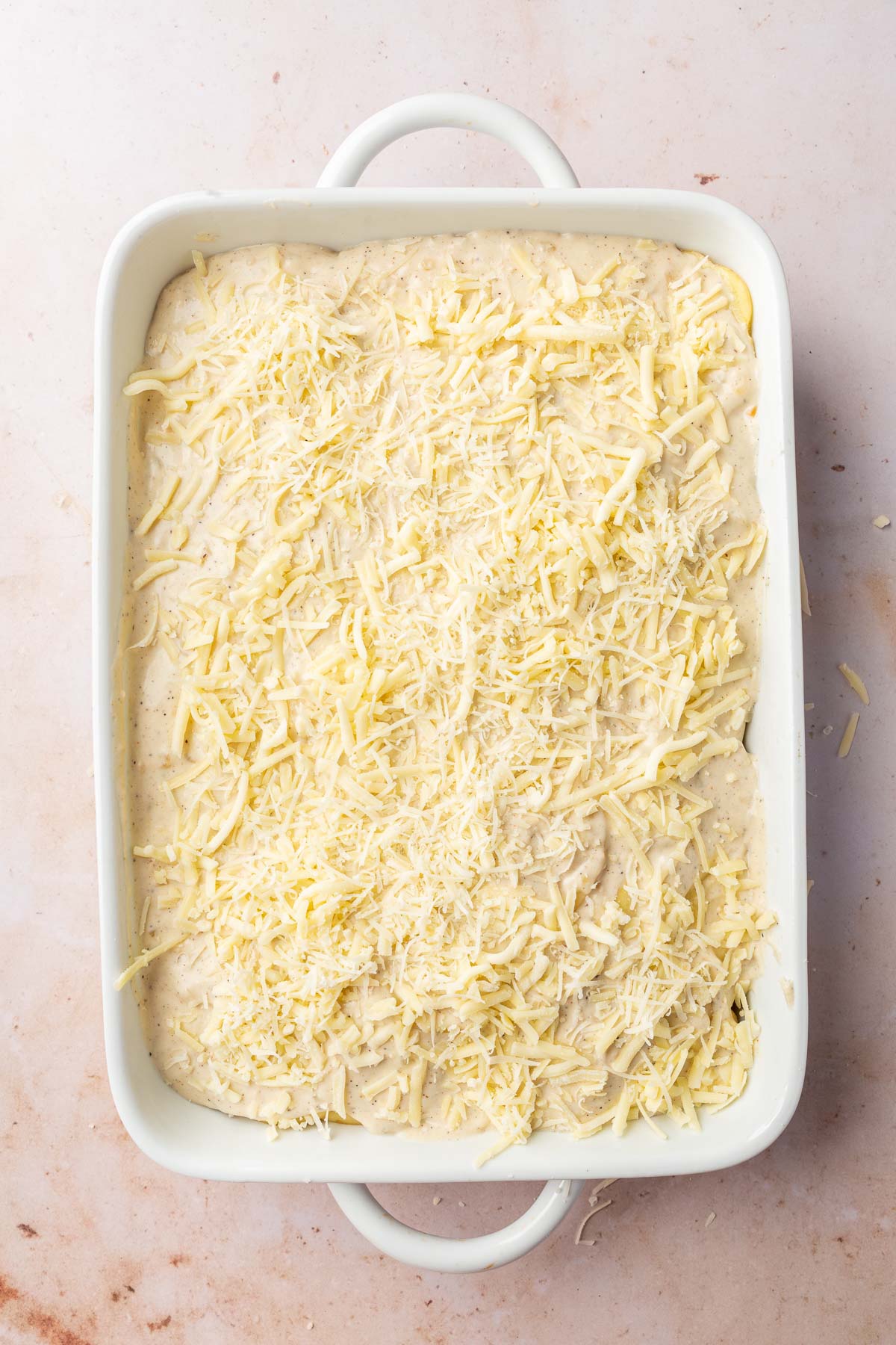 A rectangular baking dish with layers of gluten-free cream sauce topped with shredded white cheddar cheese and shredded parmesan cheese.