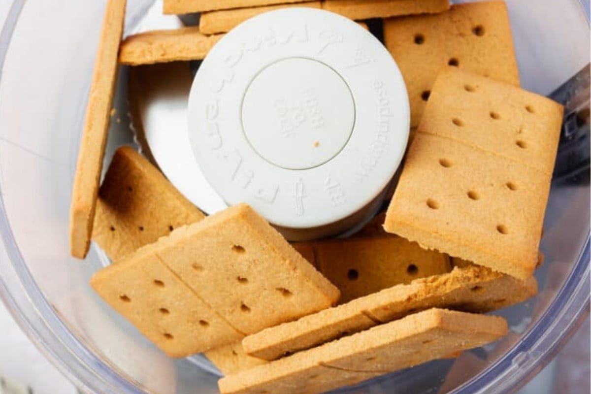 Rectangular gluten-free graham crackers are placed inside a food processor, ready to be crushed for a gluten-free graham cracker crust. The light brown crackers have small holes, and the processor blade is visible in the center.