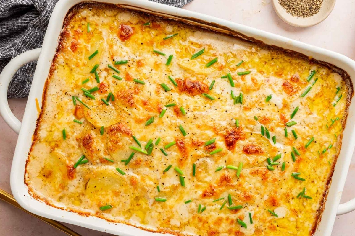 A dish of gluten-free scalloped potatoes, baked to perfection with melted cheese and chives, sits in a white rectangular dish on a light surface beside a small bowl of ground black pepper.