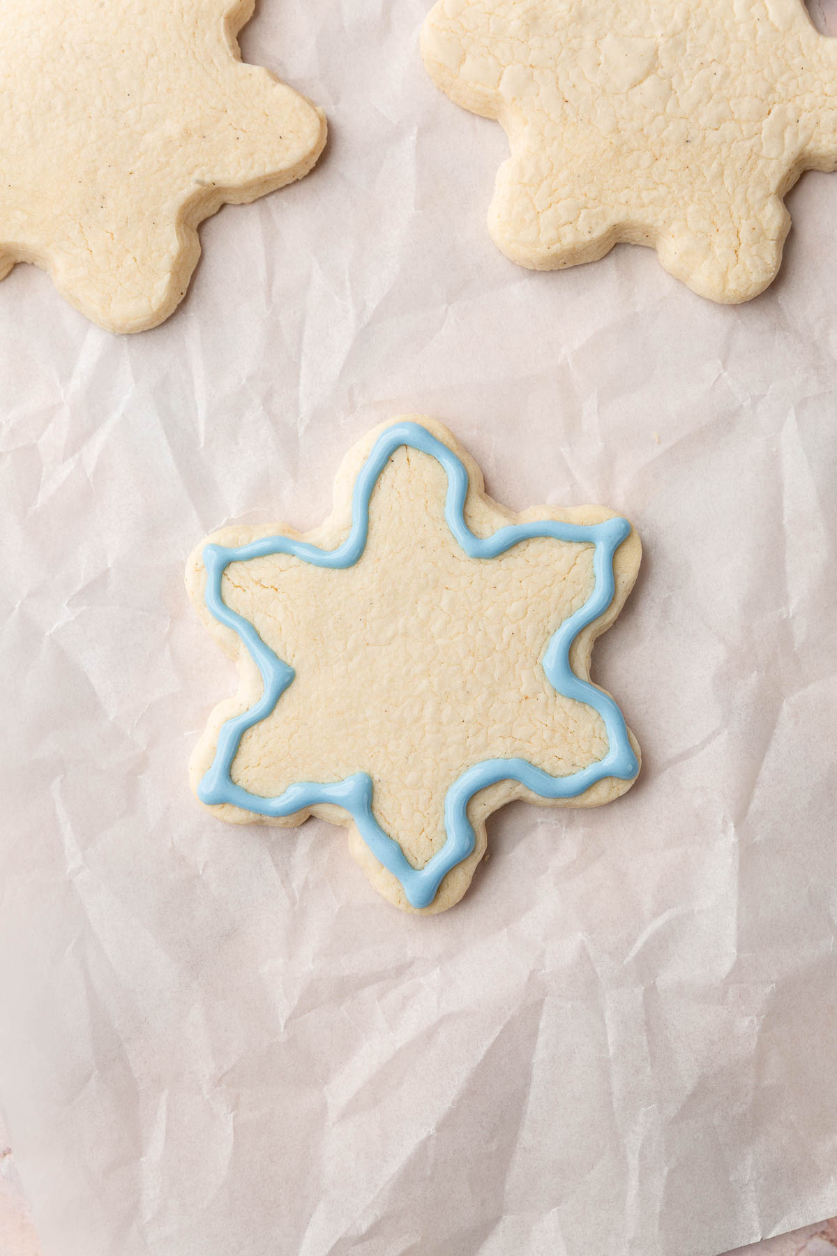 Three gluten-free snowflake cookies on a piece of parchment paper with one outlined with blue royal icing.