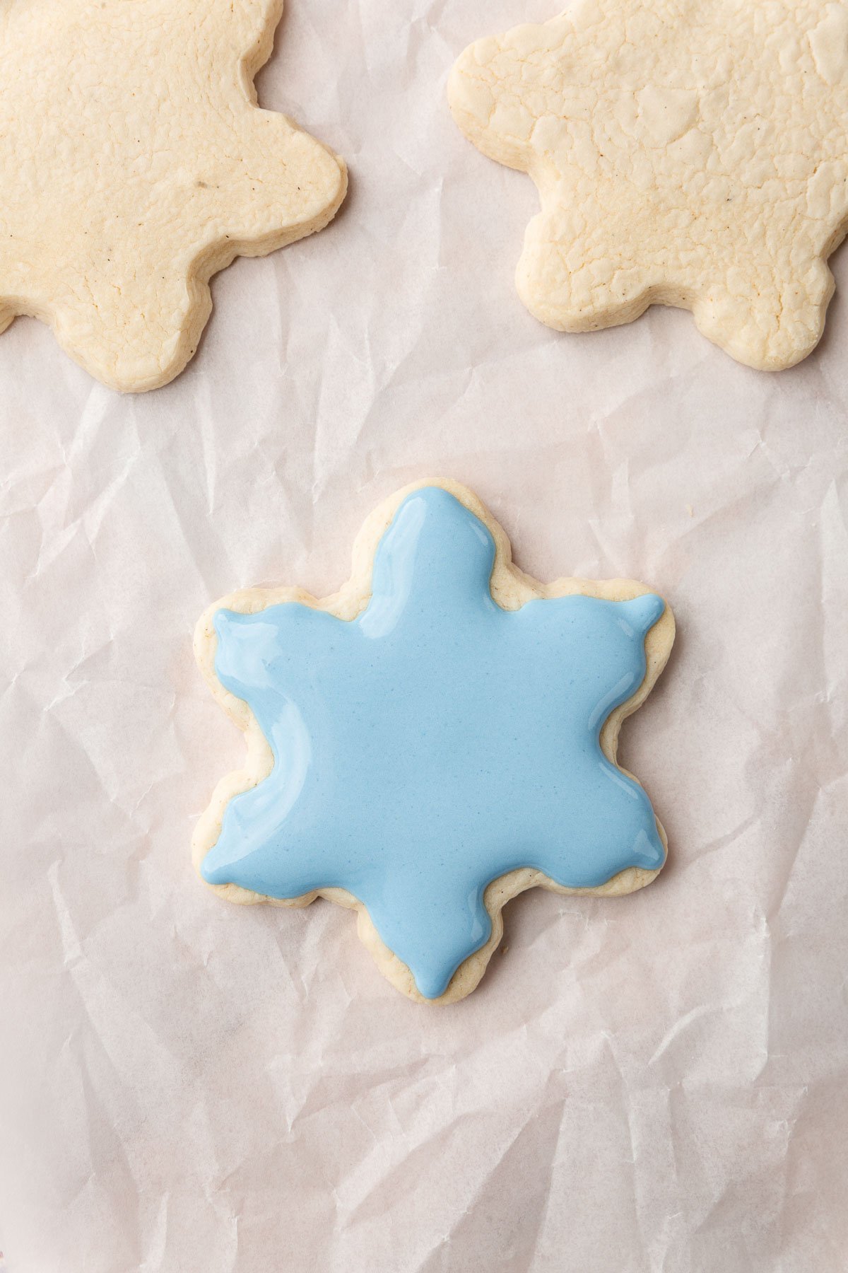 Three gluten-free snowflake cookies on a piece of parchment paper with one filled with blue royal icing.