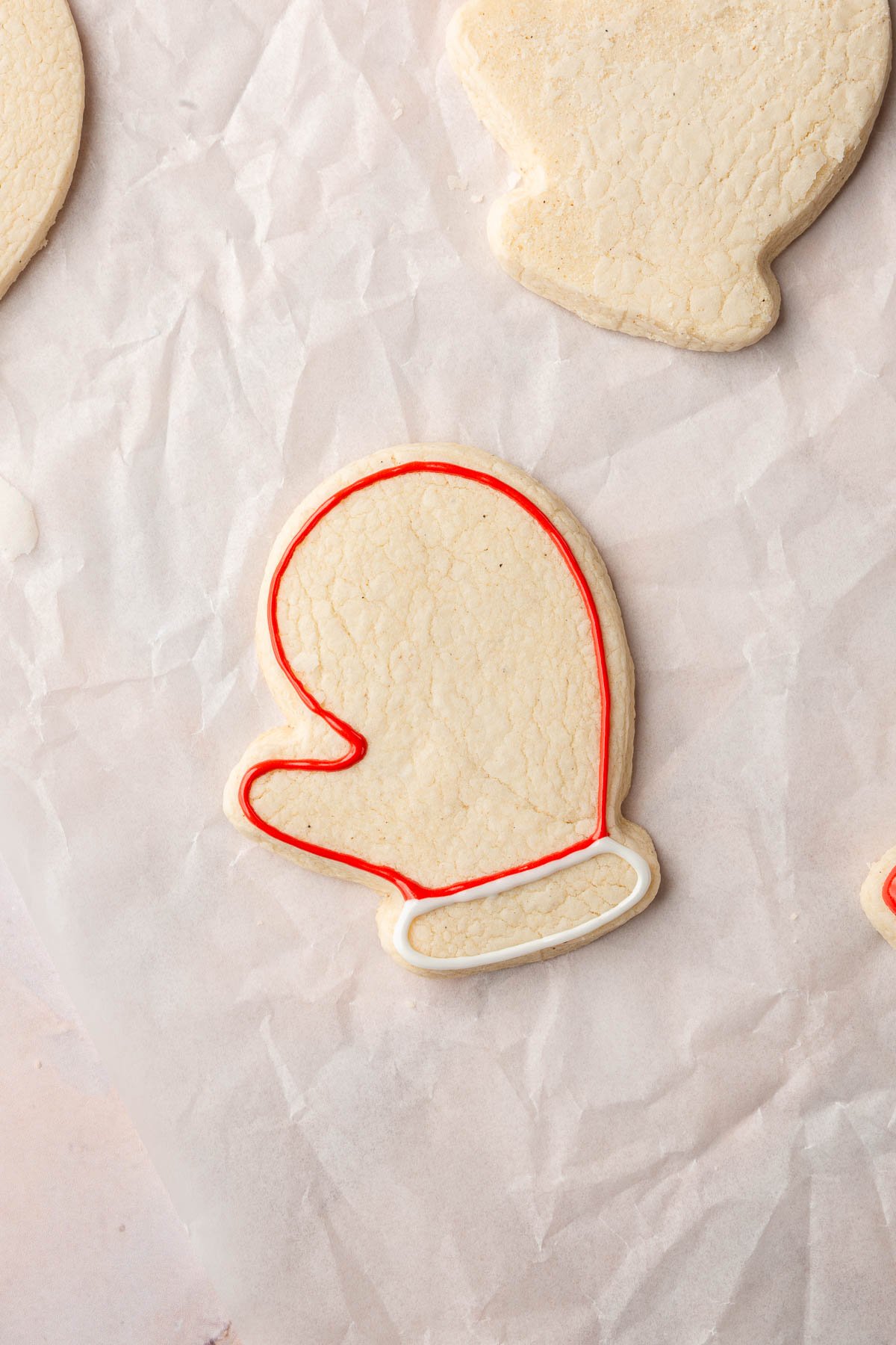 A few gluten-free mitten sugar cookies on a piece of parchment paper with one outlined with red royal icing and white royal icing.