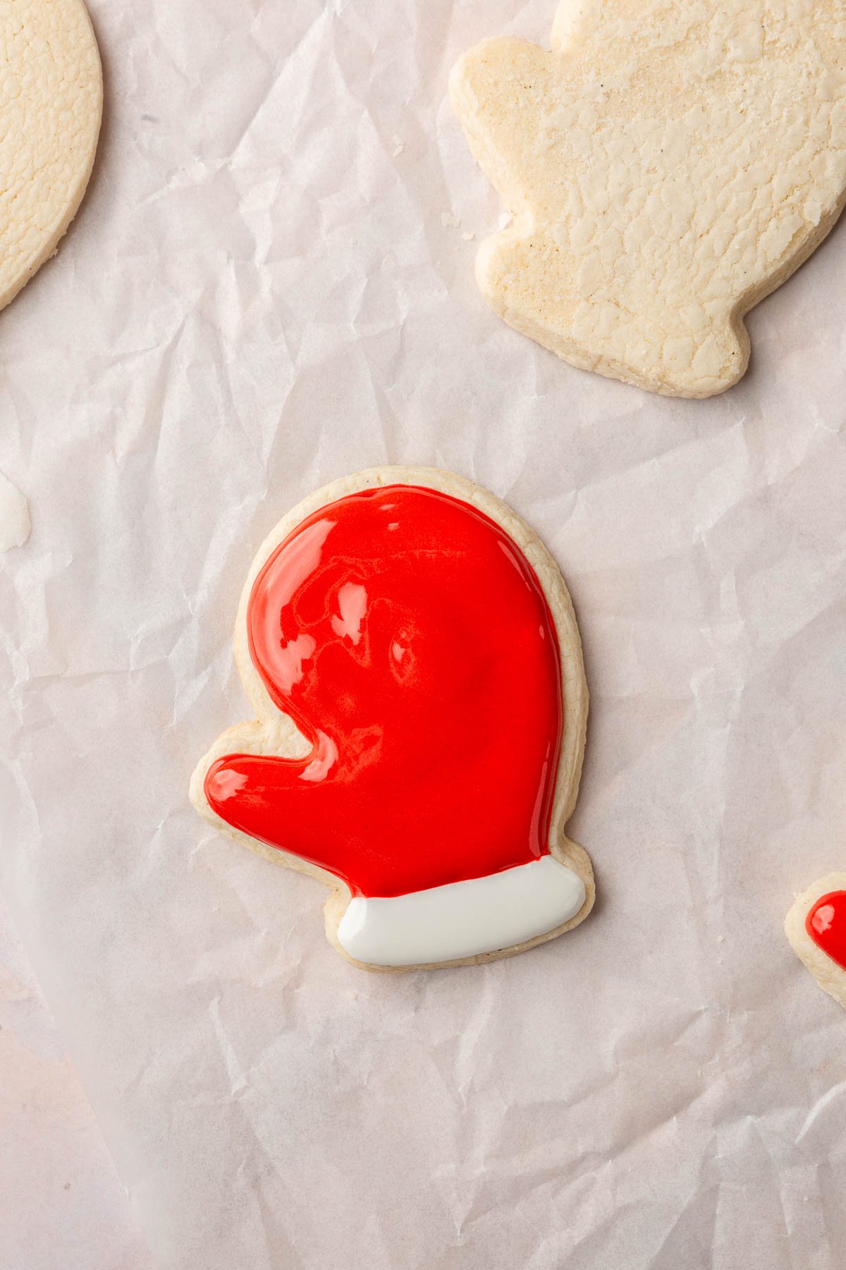 A few gluten-free mitten sugar cookies on a piece of parchment paper with one decorated with red and white royal icing.
