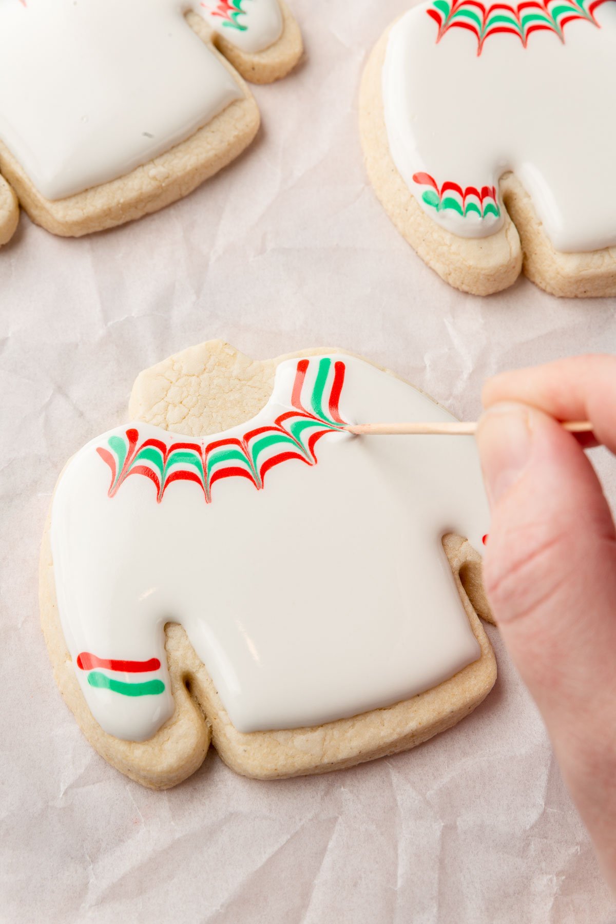 A few gluten free ugly sweater Christmas cookies on a piece of parchment paper with one decorated with white royal icing with a hand dragging a toothpick through red and green stripes to make a chevron design using a wet-on-wet technique.