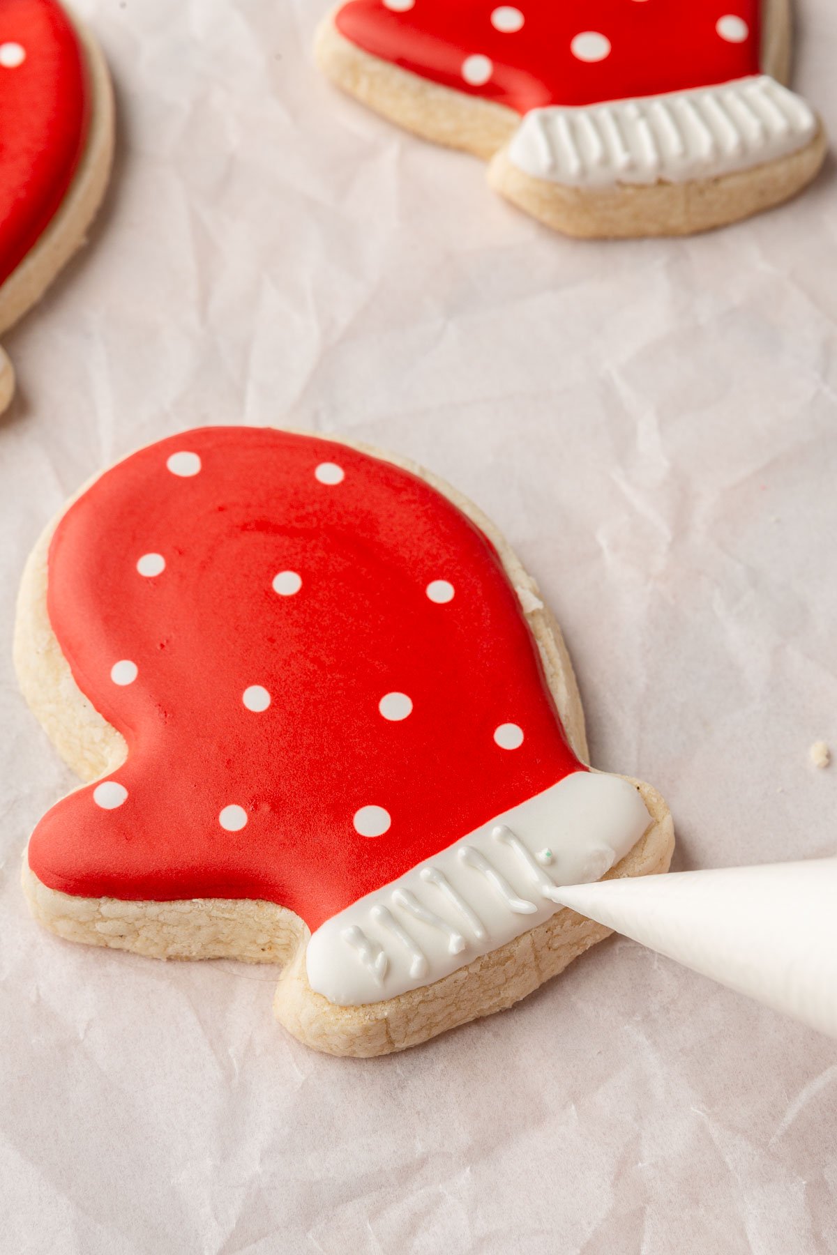 A few gluten free mitten sugar cookies with red icing and white polka dots with a bag of white royal icing making details on the cuff of the mittens.